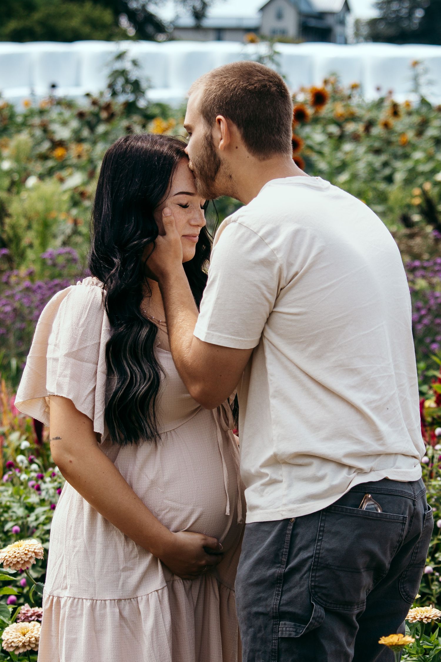 Couple sharing a tender moment in a flower garden, man kissing pregnant woman's forehead while she cradles her baby bump.
