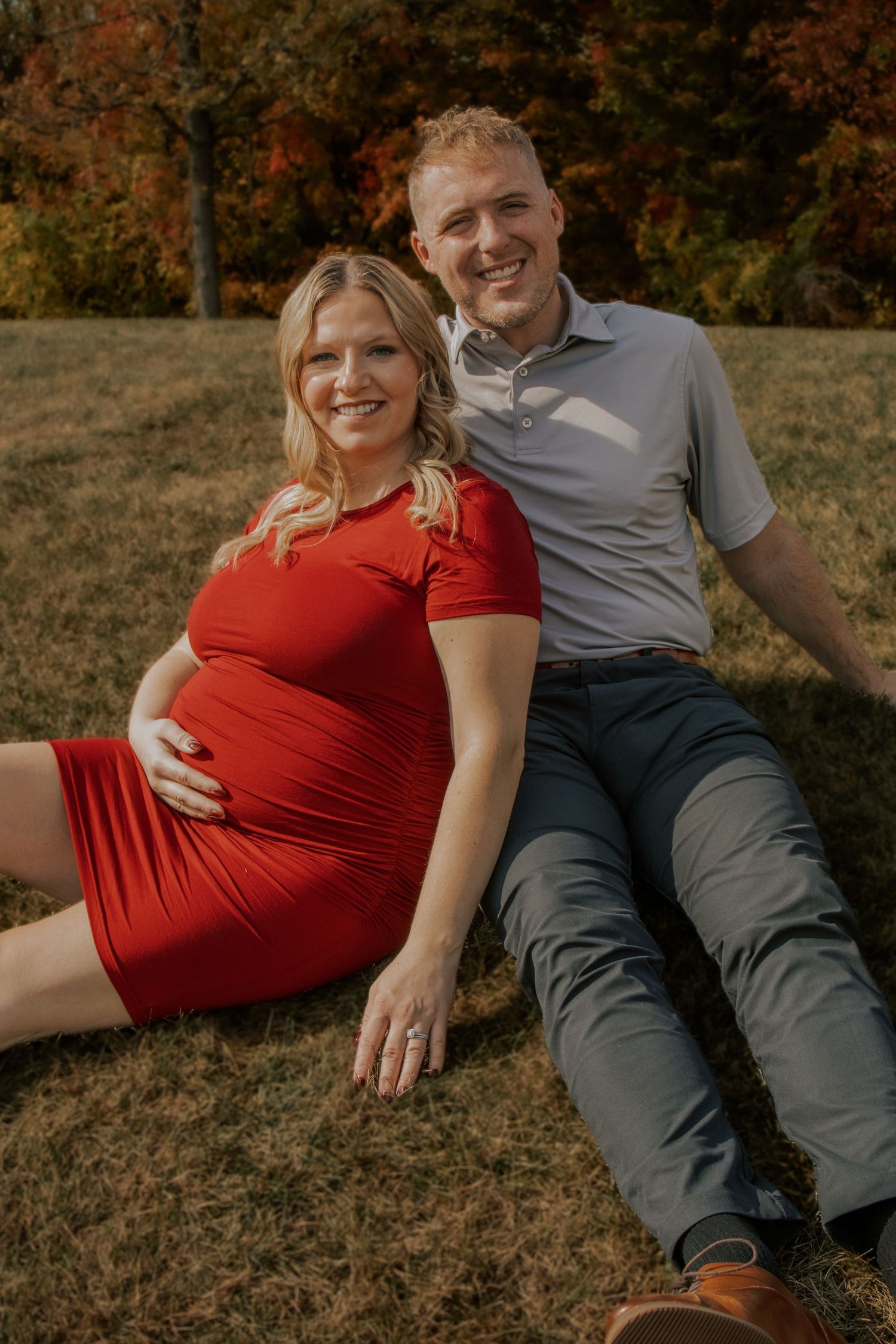Couple lounging together on autumn grass with vibrant fall foliage in background.