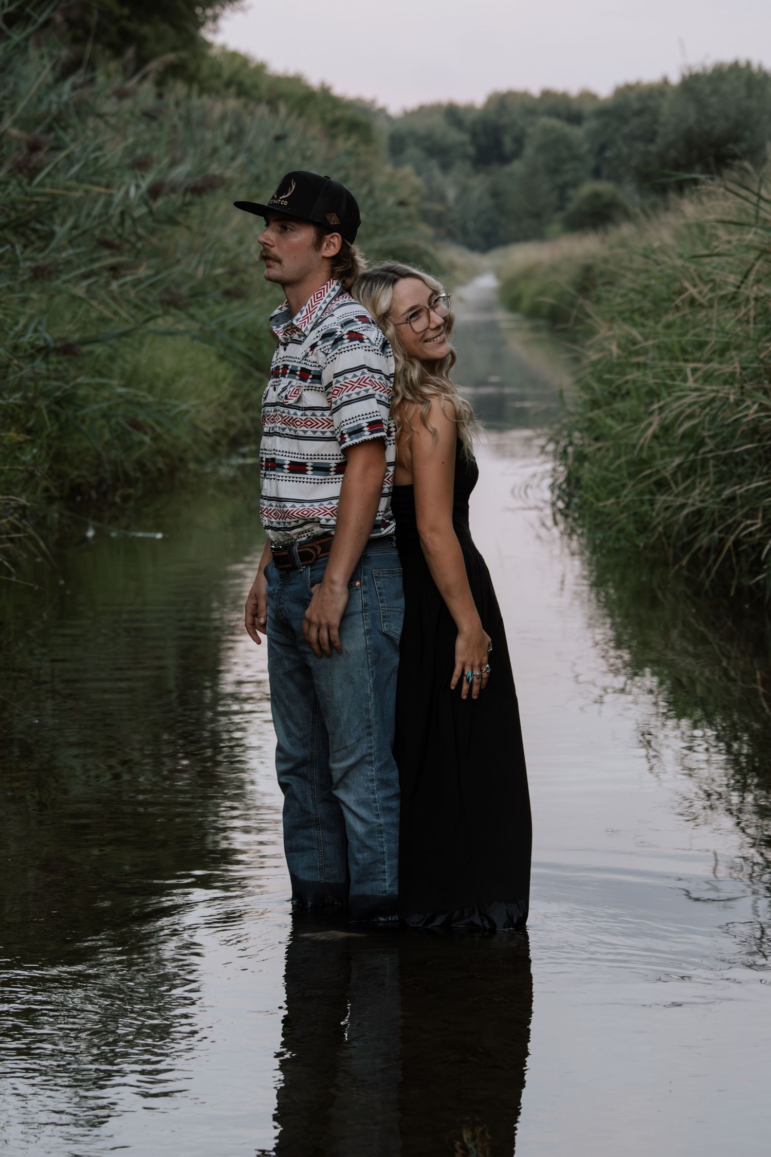 Couple standing in a shallow creek surrounded by lush greenery, man in patterned shirt and woman in black dress smiling.