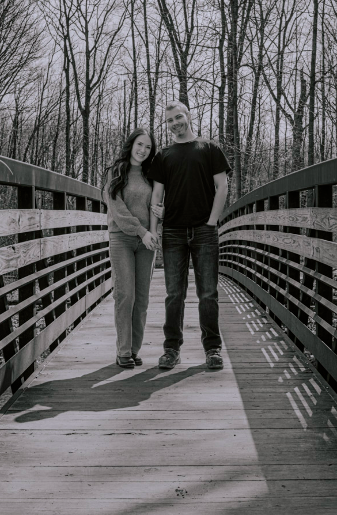 Black and white photo of a couple holding hands on a wooden bridge surrounded by bare trees.