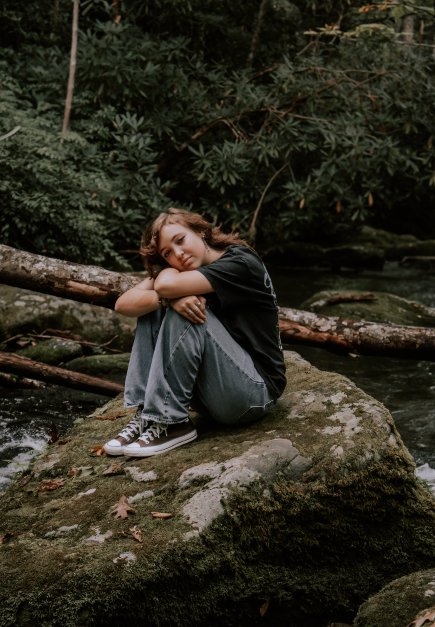 A figure rests on a mossy rock formation in a moody forest setting, showing signs of exhaustion or contemplation.
