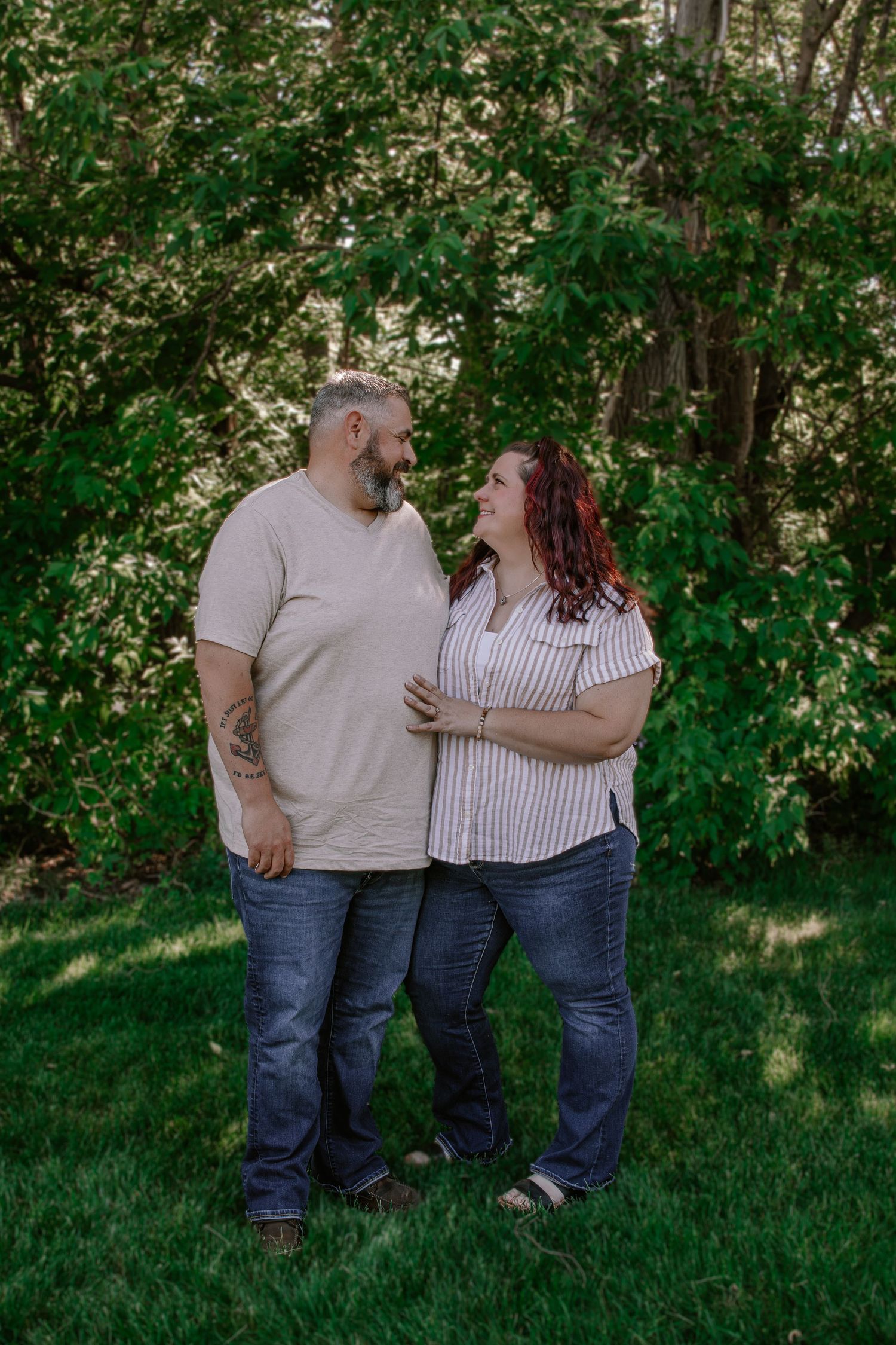 Happy couple sharing a loving glance while standing together on green grass with trees behind them.
