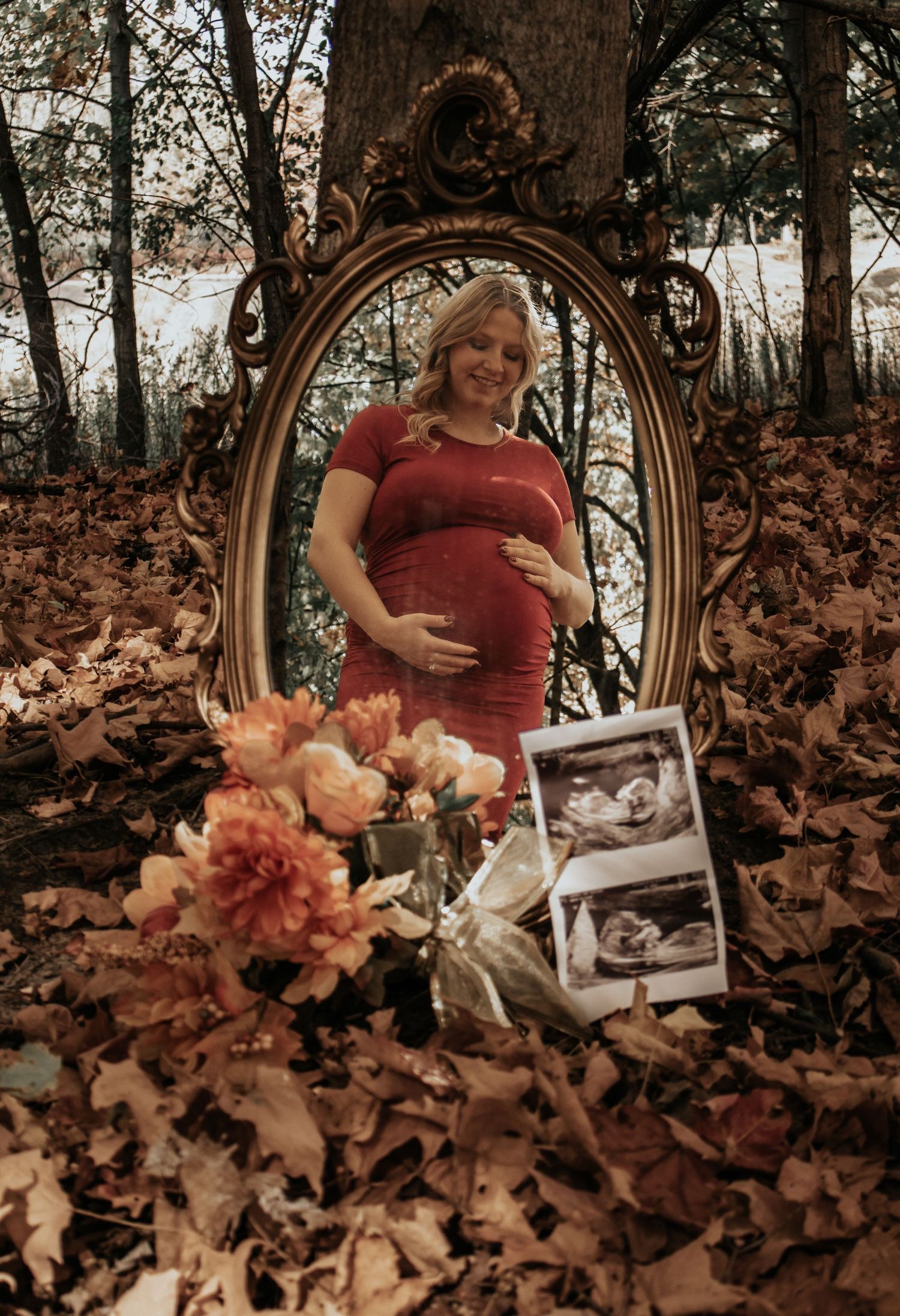 A maternity photo setup with an antique mirror, flowers, and ultrasound photos among fall leaves.
