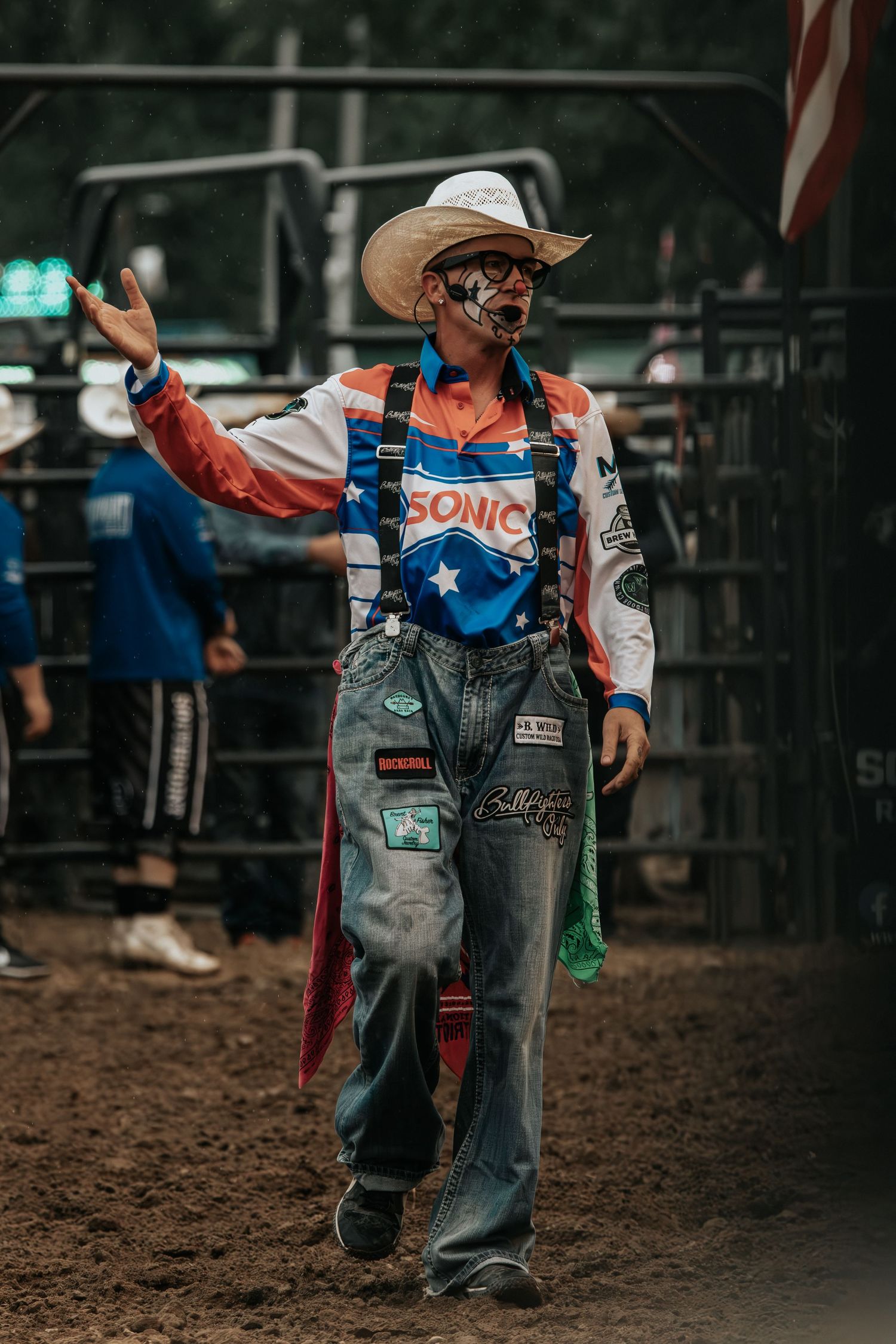 Rodeo clown in face paint, cowboy hat, Sonic-branded overalls, and American flag cape performing at a rodeo arena.