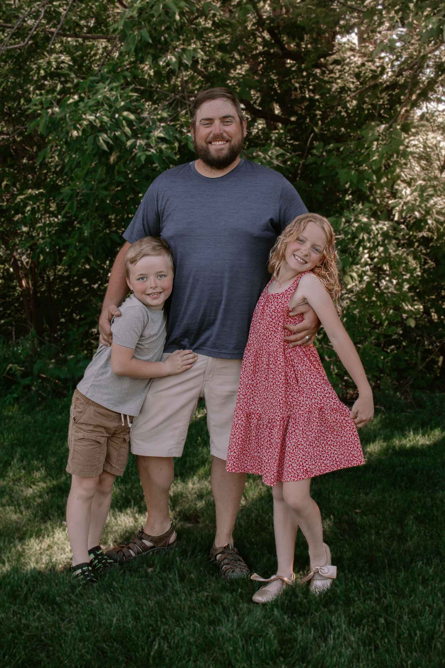 A bearded man in a blue shirt poses outdoors with two smiling children in a lush green backyard.