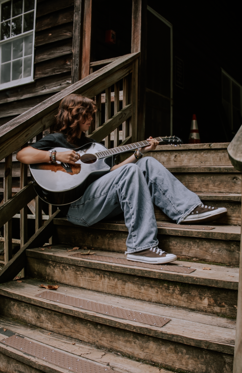 A person sits on wooden steps playing an acoustic guitar while wearing jeans and sneakers.