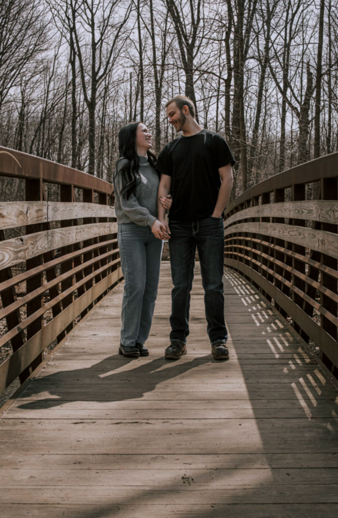 Couple holding hands and smiling at each other on a wooden bridge surrounded by bare trees.