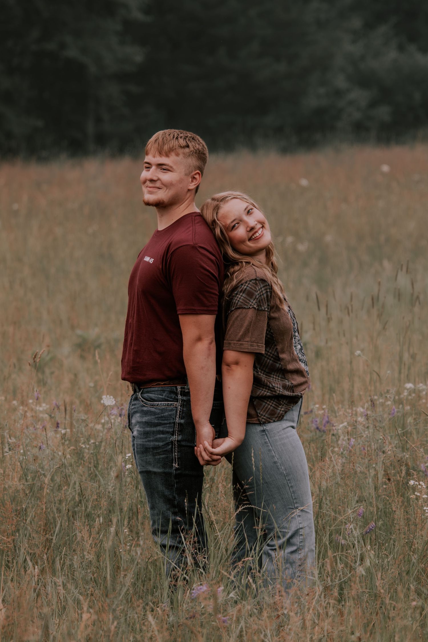 A young couple holds hands in a golden meadow, the woman leaning on the man's back, both smiling warmly.