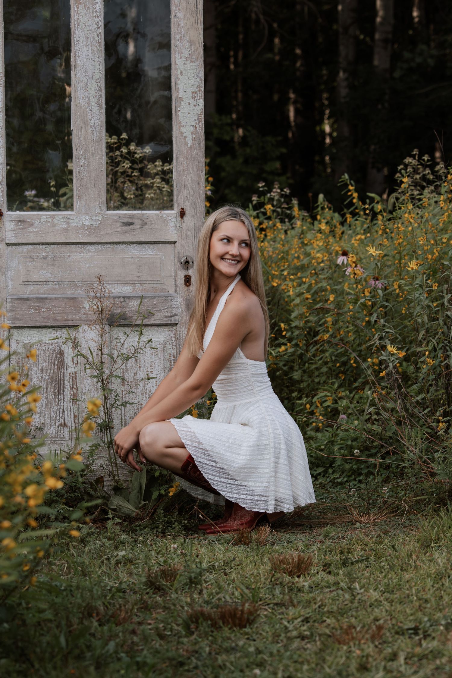 Young blonde woman in white dress and cowboy boots crouching beside rustic wooden door surrounded by wildflowers.