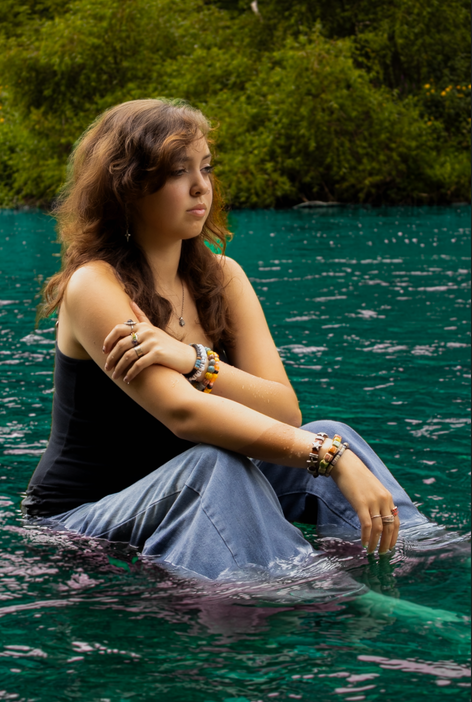 A dreamy portrait of someone sitting in turquoise waters surrounded by lush greenery wearing a black top and flowing skirt.