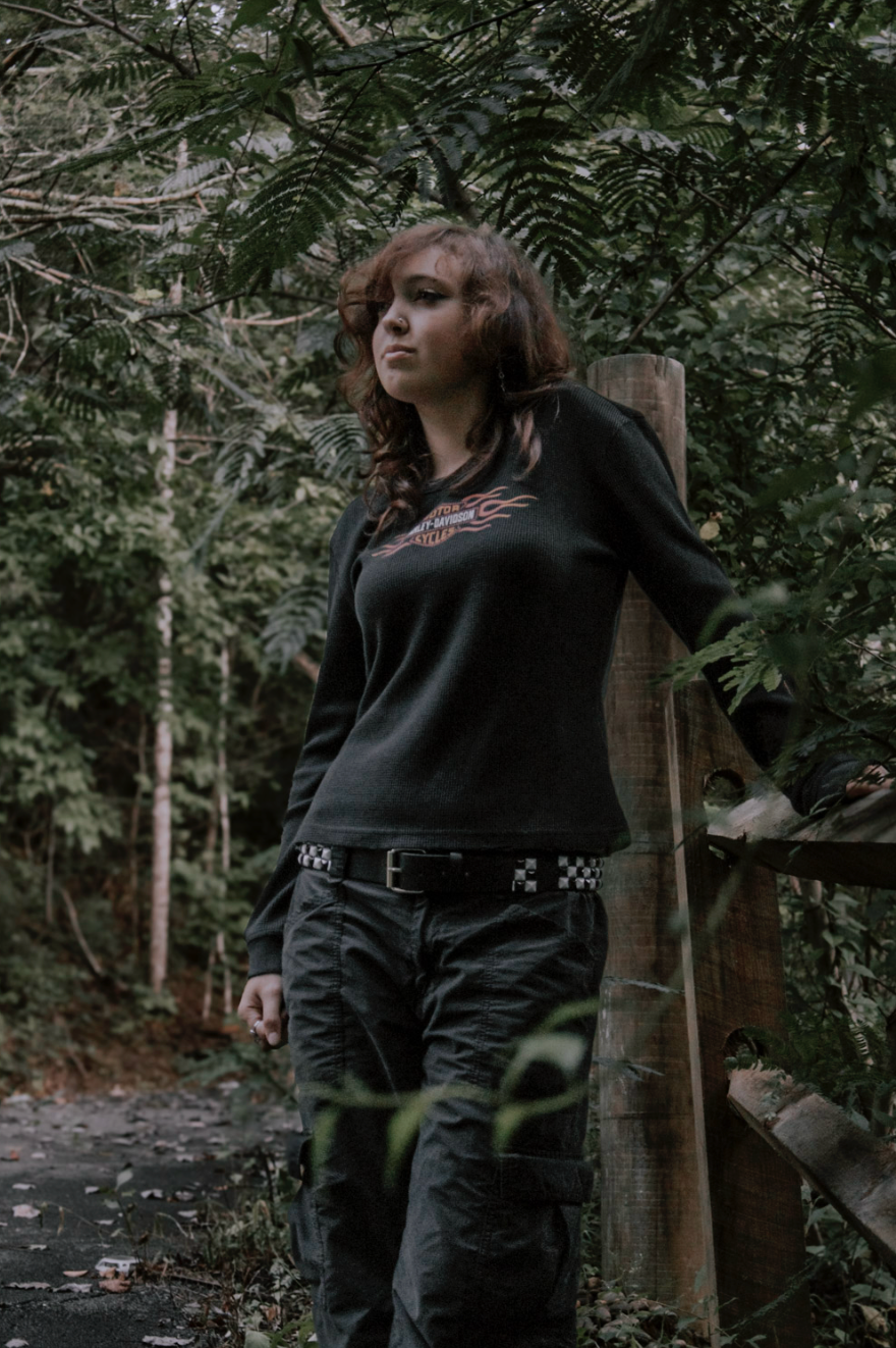Person in dark clothing poses against wooden post in atmospheric forest setting with dramatic lighting.