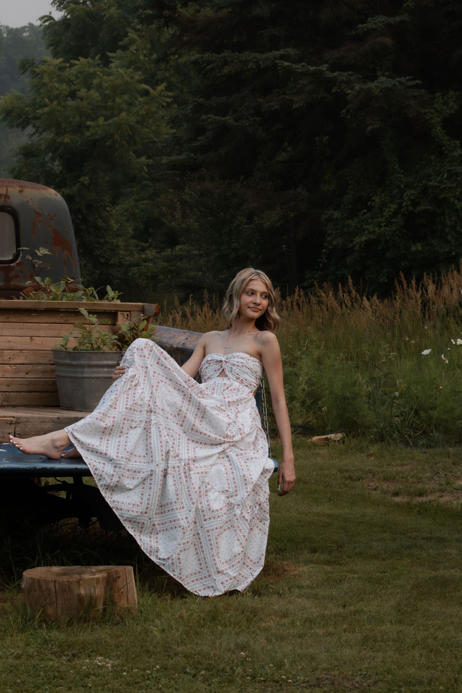A young woman in a flowing white strapless dress sits on a vintage truck tailgate surrounded by lush greenery.