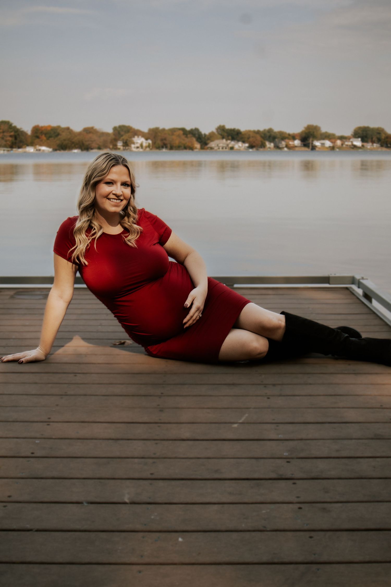 A maternity photo session on a wooden dock with a lake and autumn trees in the background.