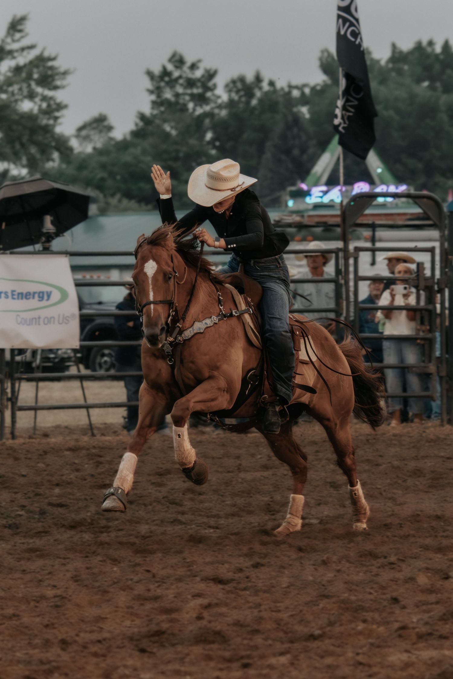 Cowgirl in white hat rides a chestnut horse at a rodeo competition, raising her hand while galloping on dirt arena.
