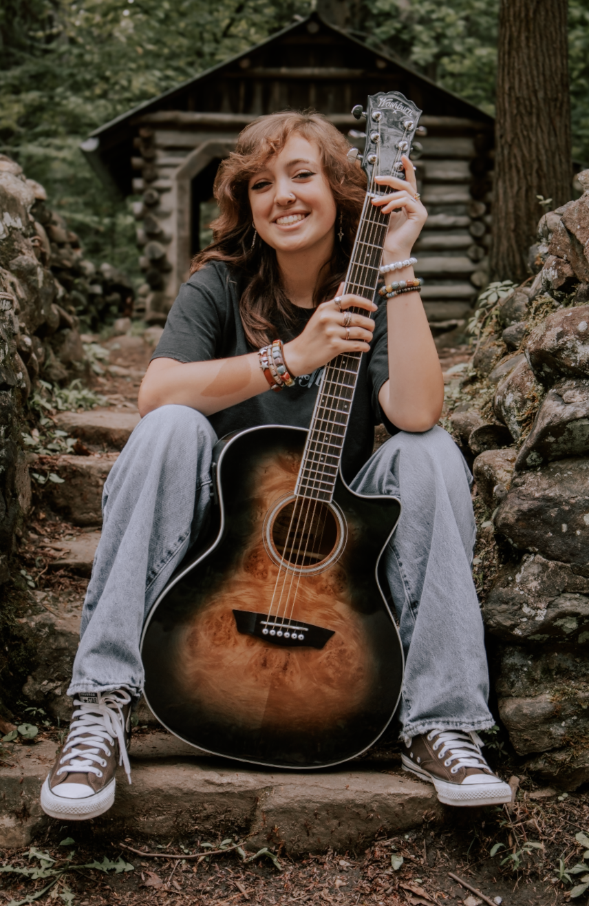 Acoustic guitarist in jeans sits on rustic woodland rocks with sunburst guitar in casual outdoor portrait session.