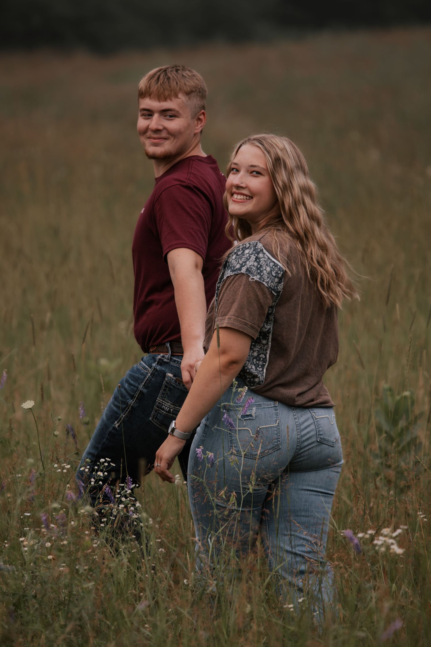 Couple holding hands and smiling in a wildflower field, wearing jeans and casual tops.
