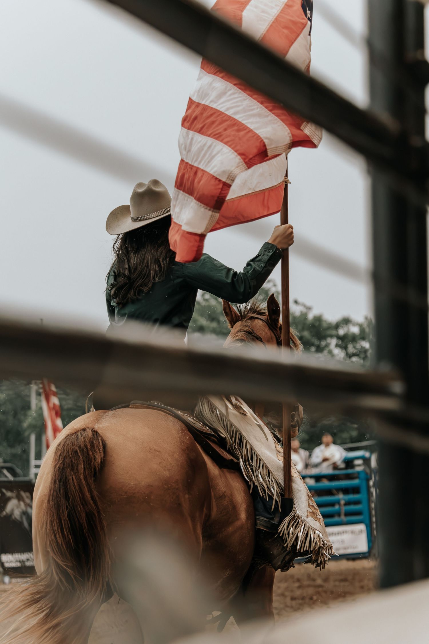 Cowgirl on horseback carrying American flag at rodeo arena, viewed through fence rails.