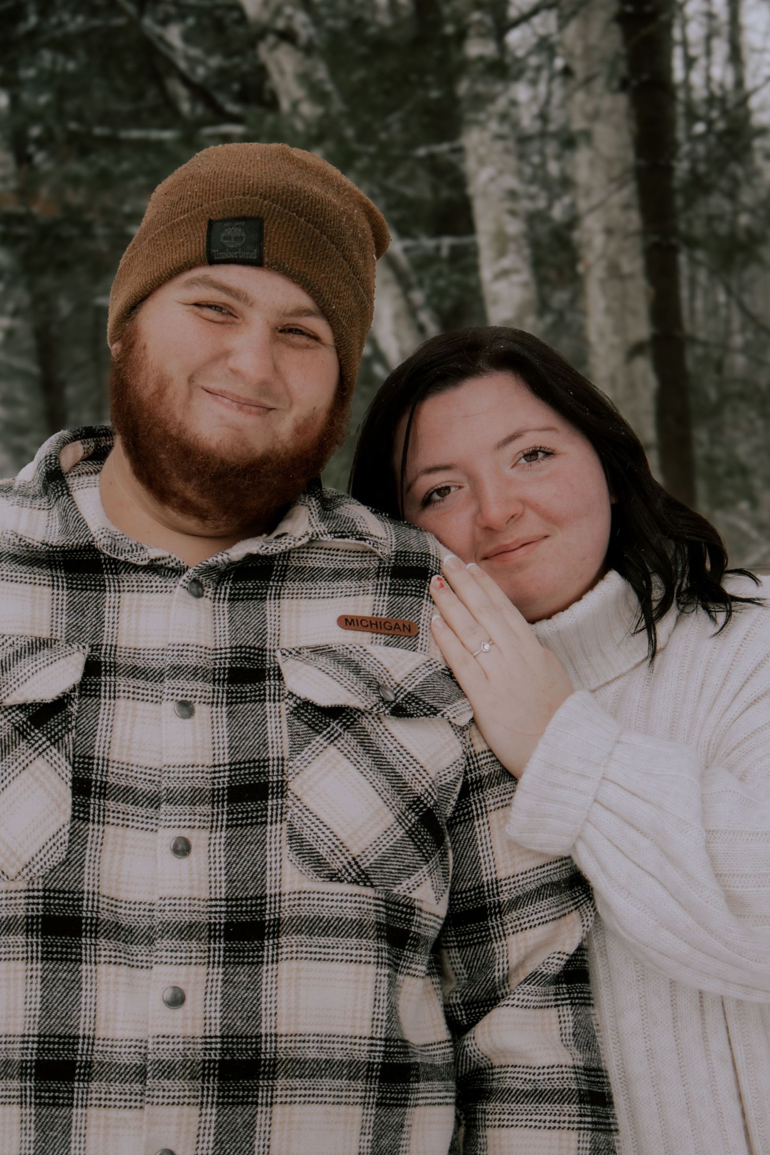 A couple wearing plaid and white clothing pose together in a winter forest setting.