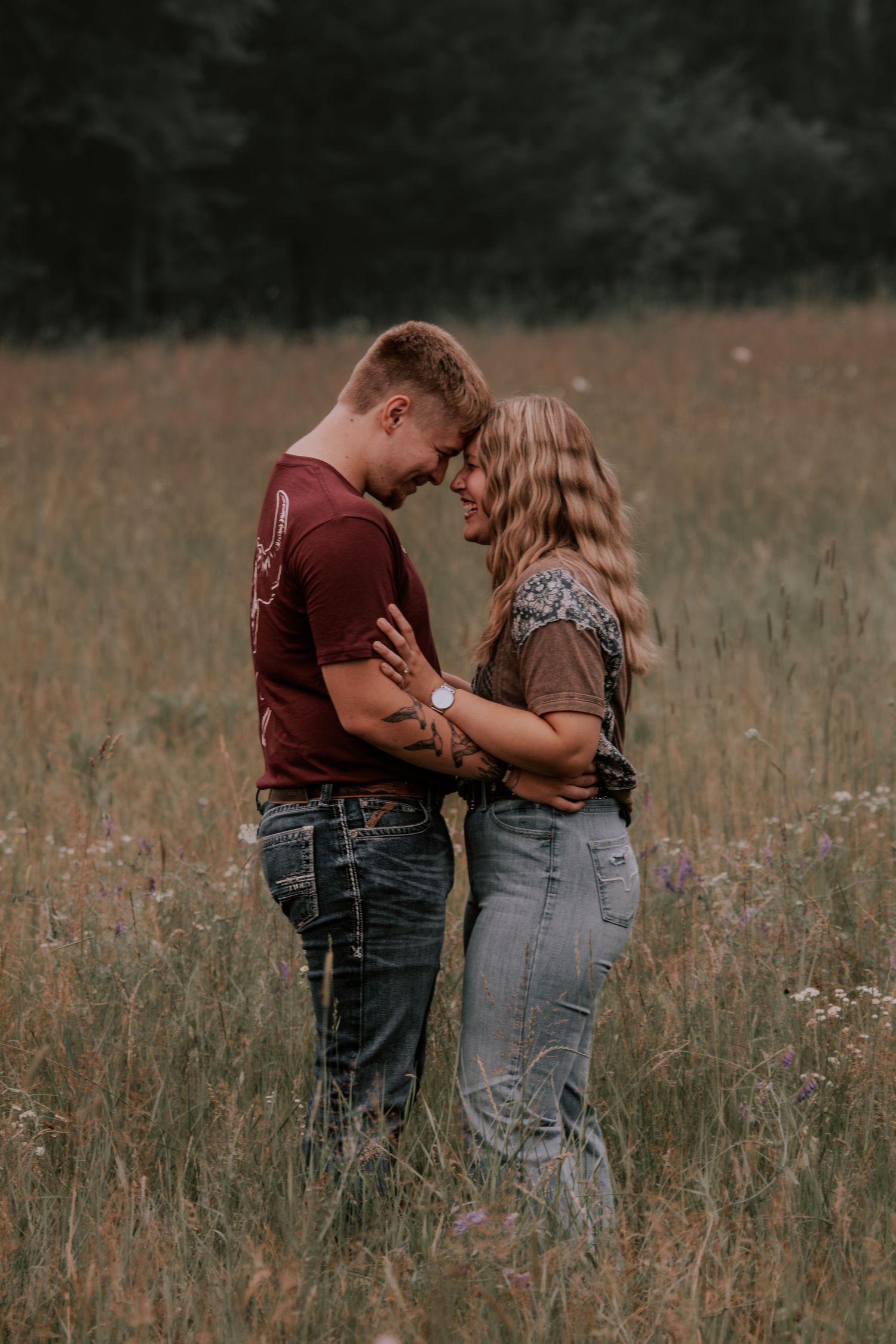 A couple embraces forehead-to-forehead in a golden meadow, wearing casual jeans and tops in a romantic outdoor portrait.