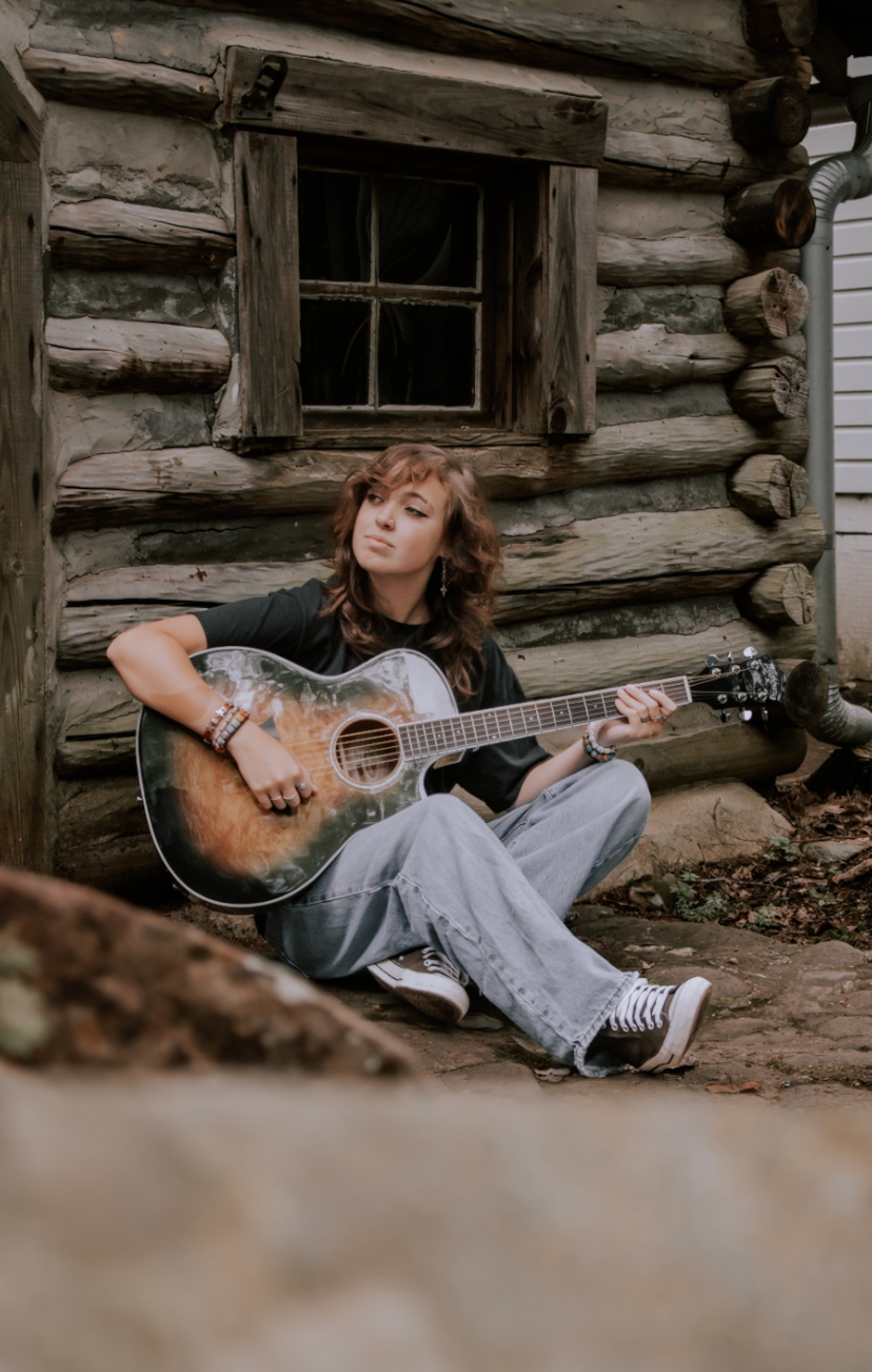 A rustic scene with someone playing acoustic guitar against a log cabin wall in warm natural light.