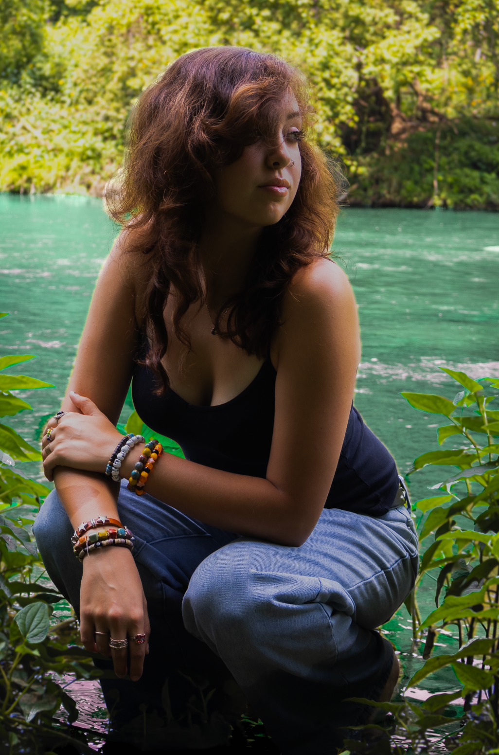 Someone wearing colorful bracelets poses thoughtfully by a vibrant turquoise lake with green foliage in the background.
