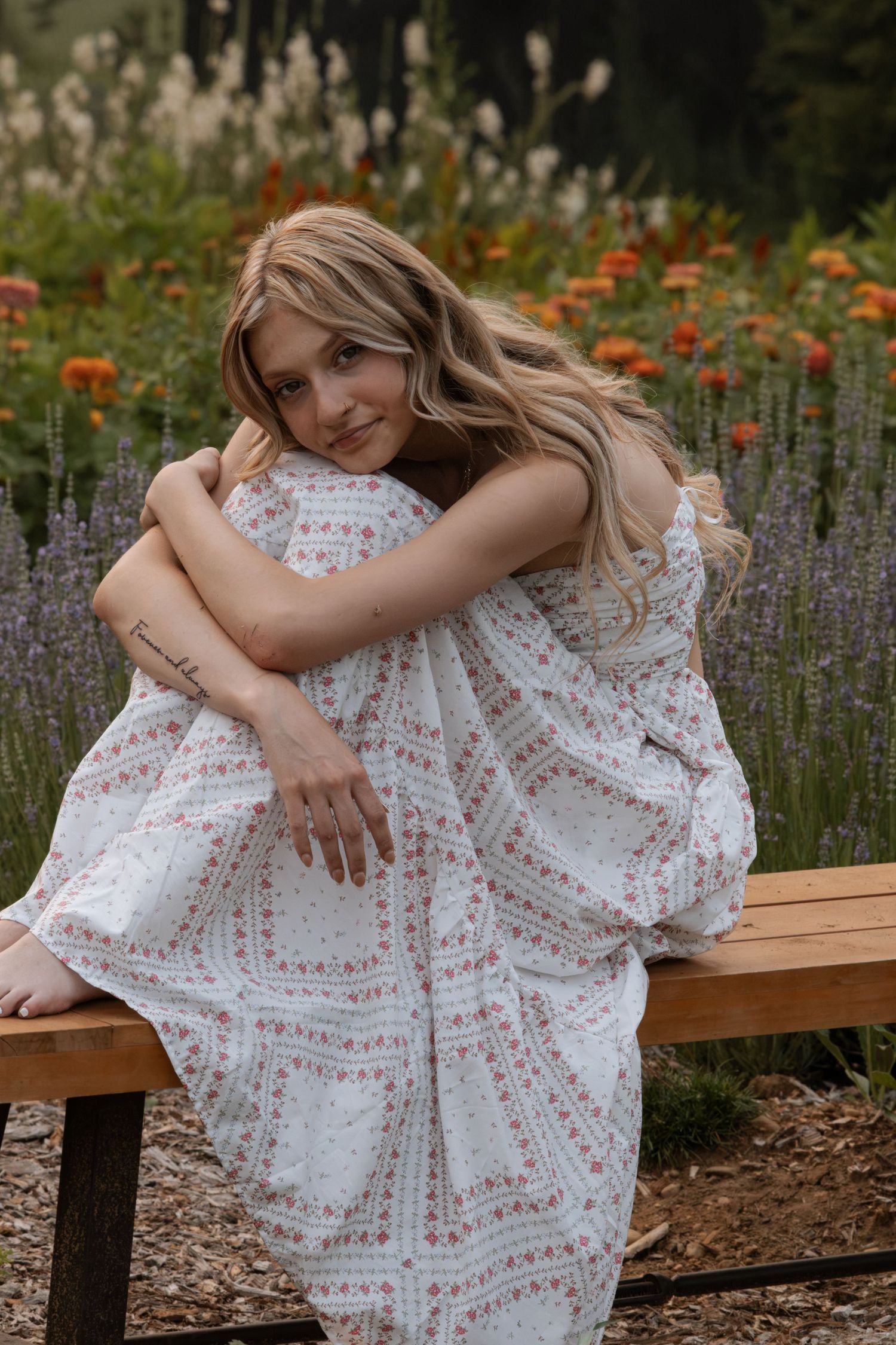 Young woman in floral white dress sitting on wooden bench, hugging knees, surrounded by lavender and orange wildflowers.