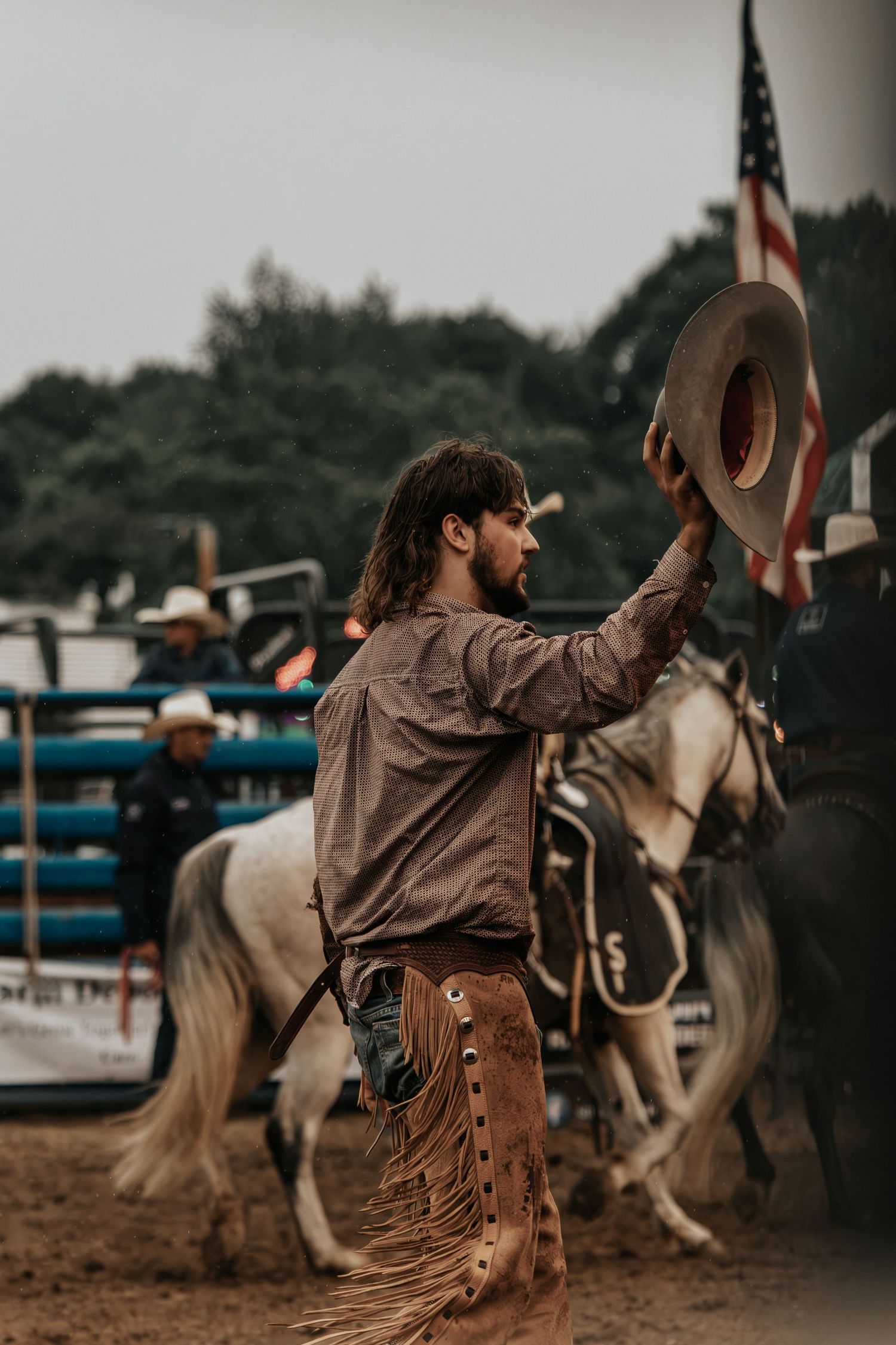 A cowboy in fringed chaps raises his hat at a rodeo with an American flag and horse in the background.
