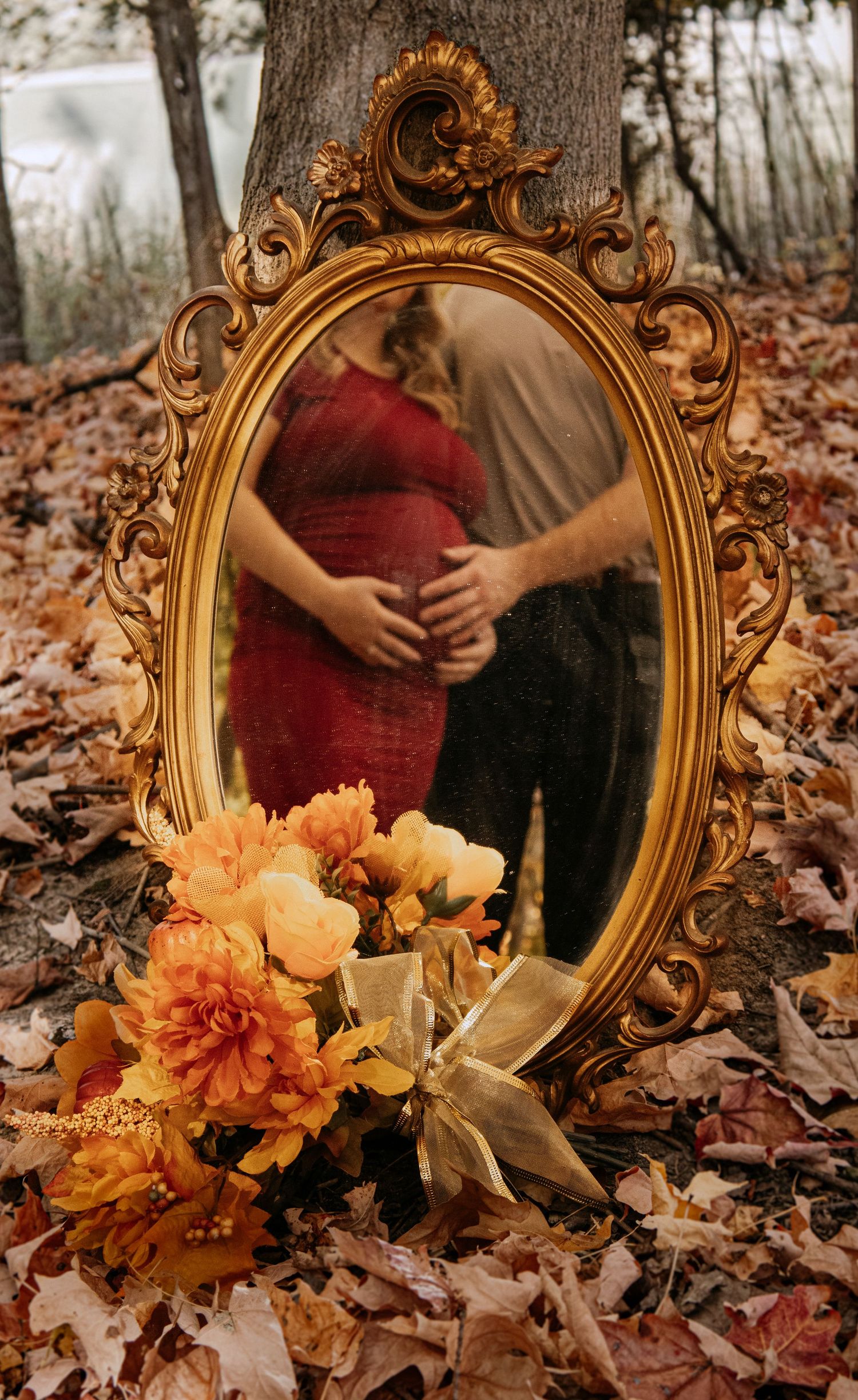 Ornate gold oval mirror reflecting pregnant couple embracing outdoors, surrounded by autumn leaves and orange flowers.