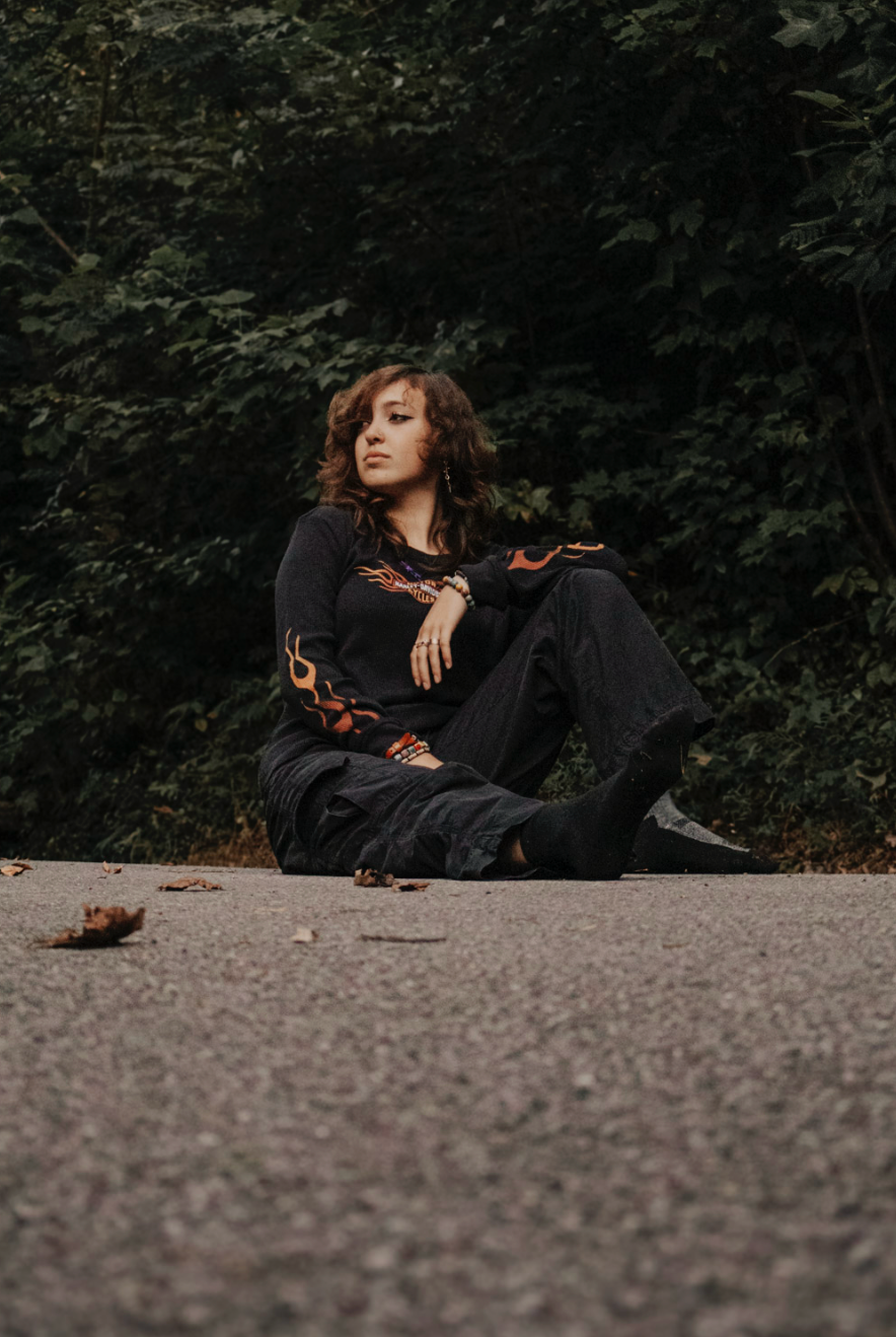A figure in a black dress reclines dramatically on a gravel path surrounded by dark foliage.