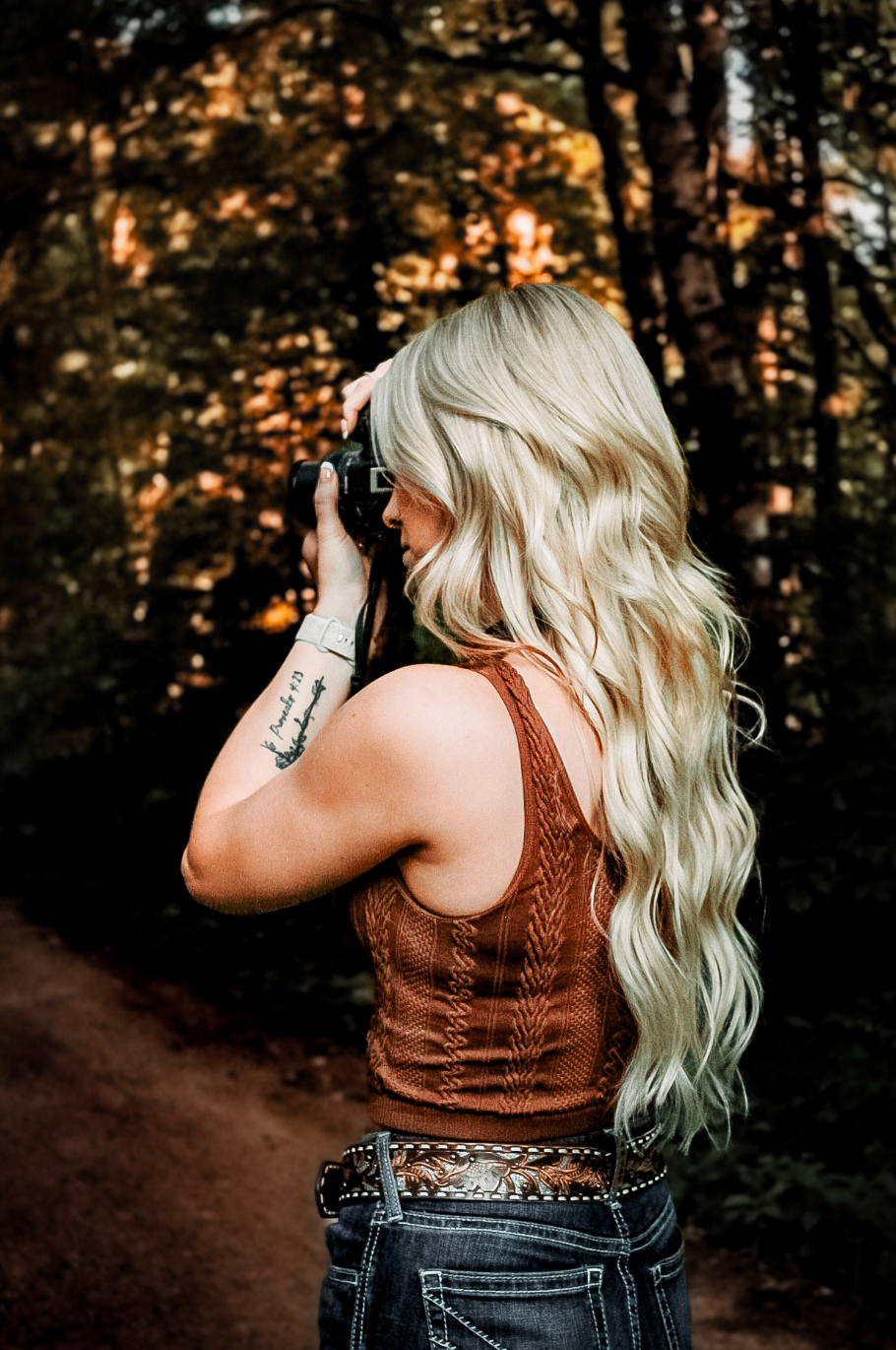 A photographer with long blonde wavy hair wearing a brown tank top takes photos in an autumn forest at sunset.