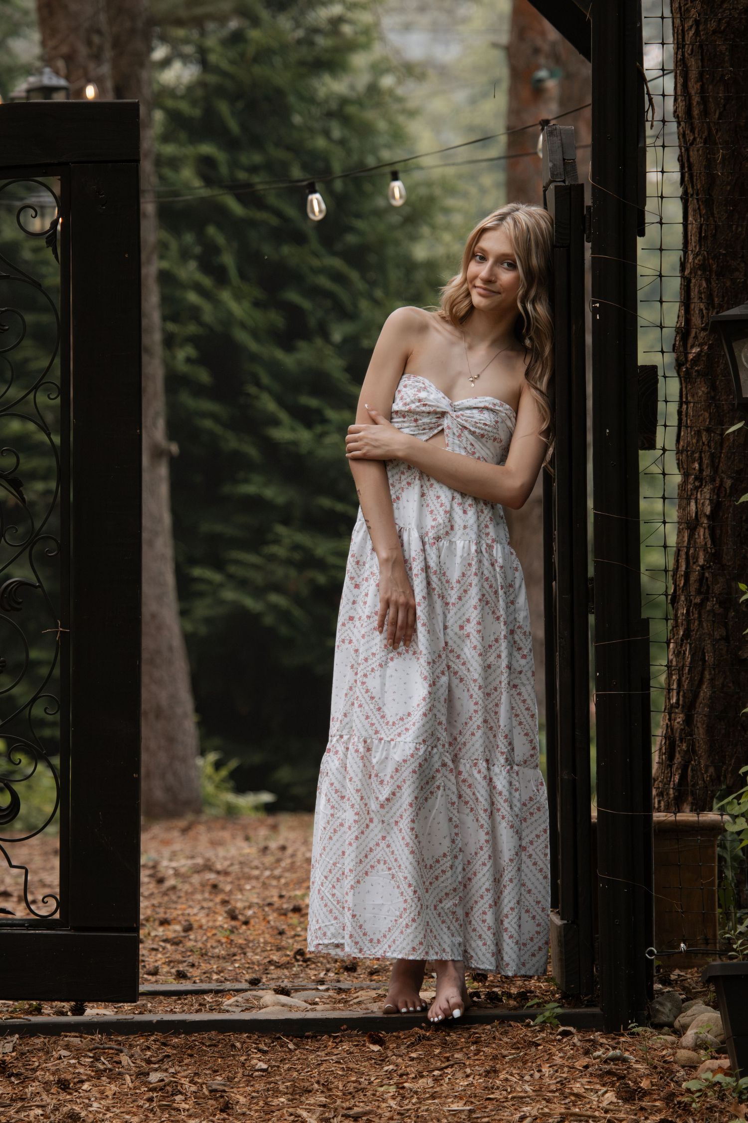 Woman in strapless floral maxi dress standing in garden gate surrounded by tall trees and string lights.