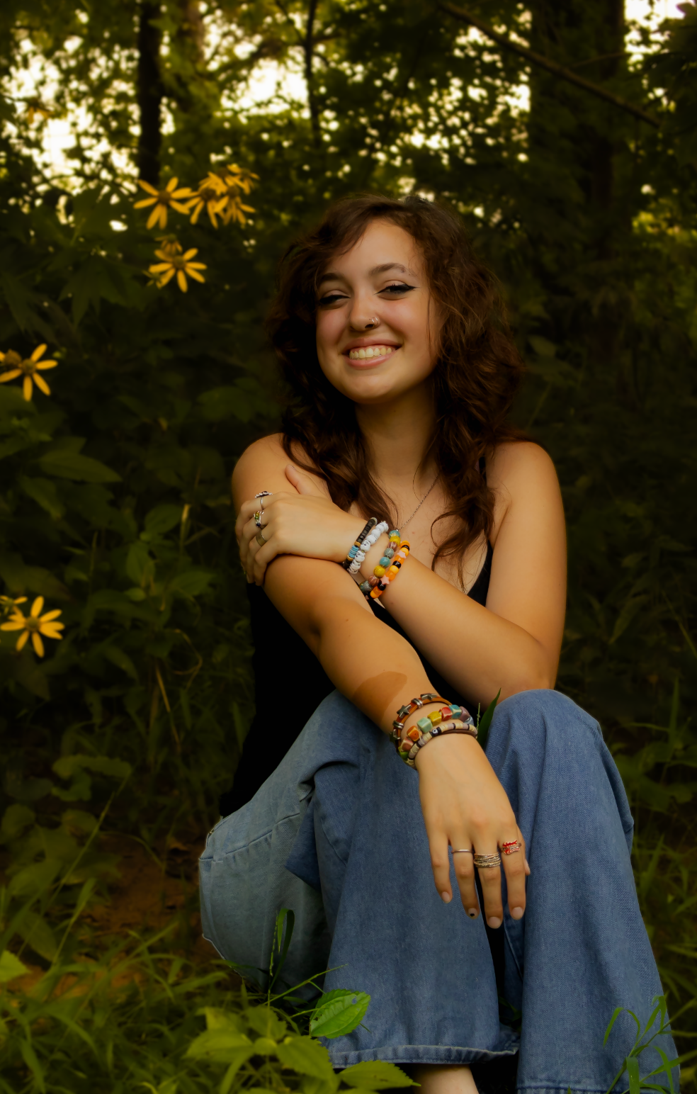 A curly-haired person sits in blue jeans among wildflowers during golden hour lighting in a garden setting.