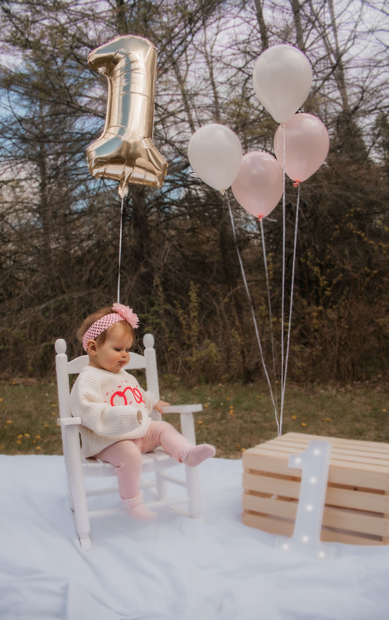 Baby girl in pink sits in white chair with gold number 1 balloon and pink balloons for first birthday photo.