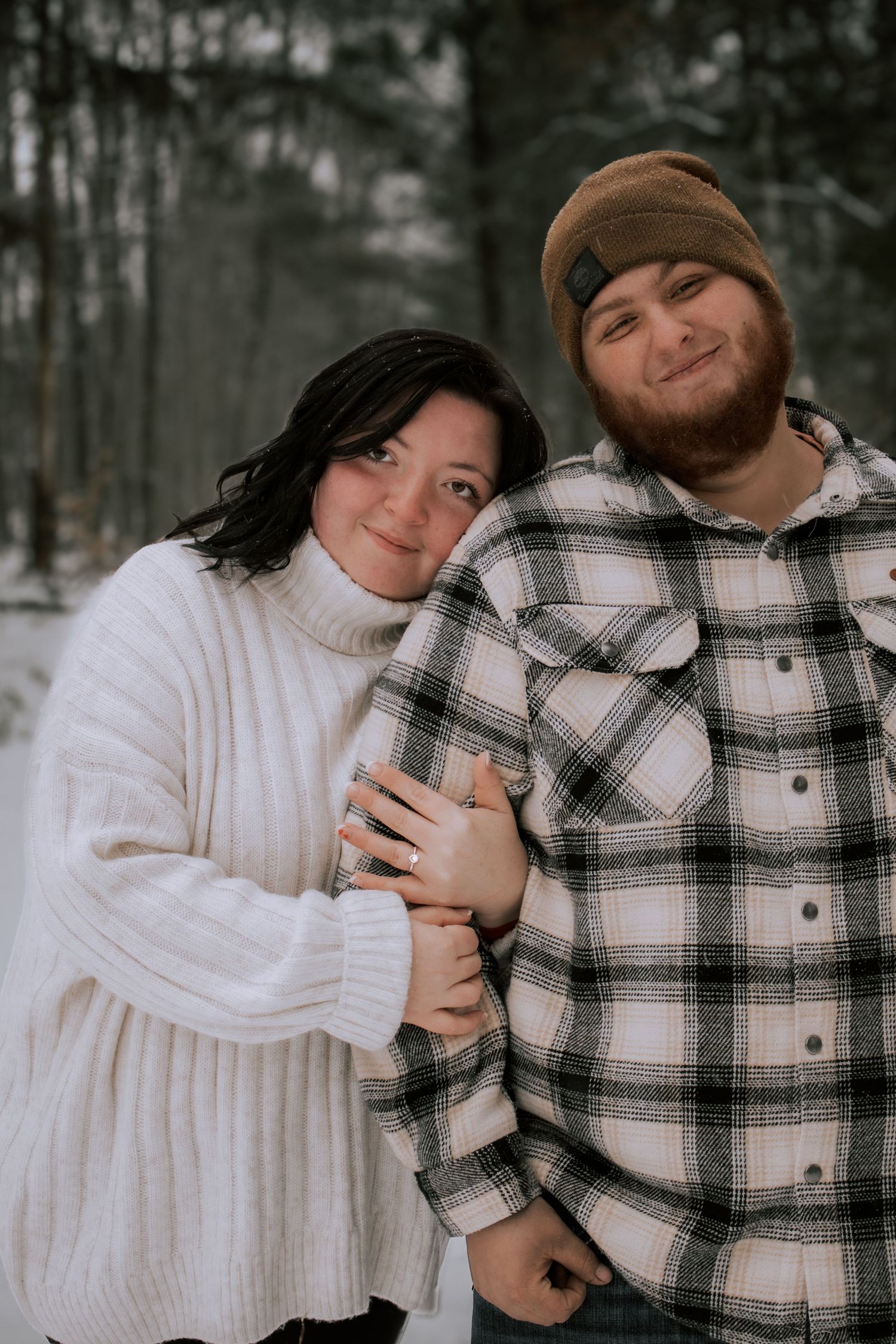 Couple embracing in snowy winter forest, woman in white sweater leaning on man in plaid flannel shirt and beanie.