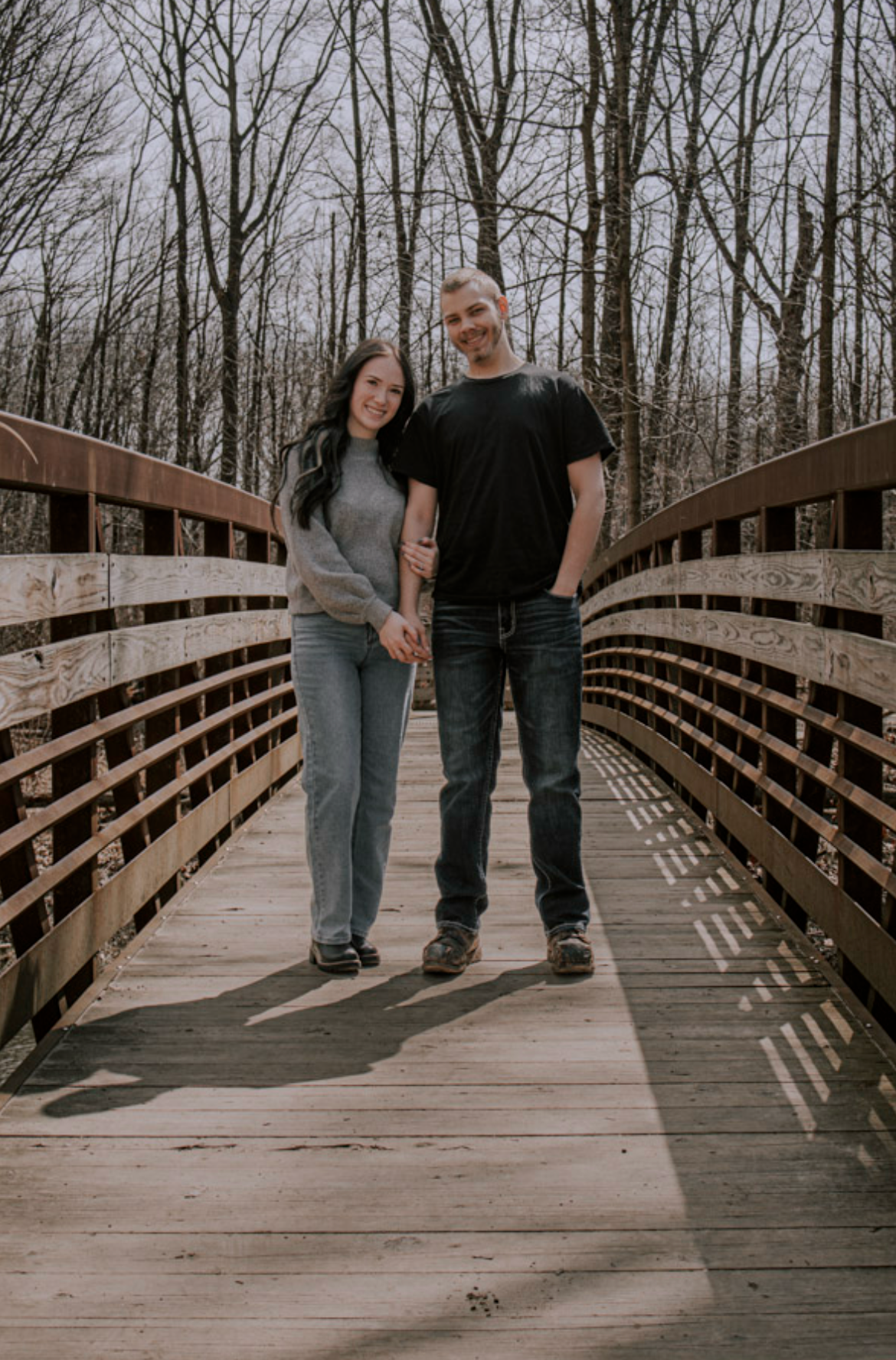 Couple holding hands and smiling on a wooden bridge surrounded by bare trees in a park.