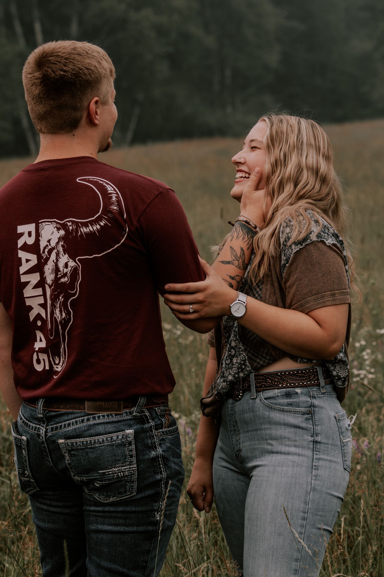 A laughing blonde woman in jeans and a brown top stands with a man wearing a maroon Rank 45 t-shirt in a field.
