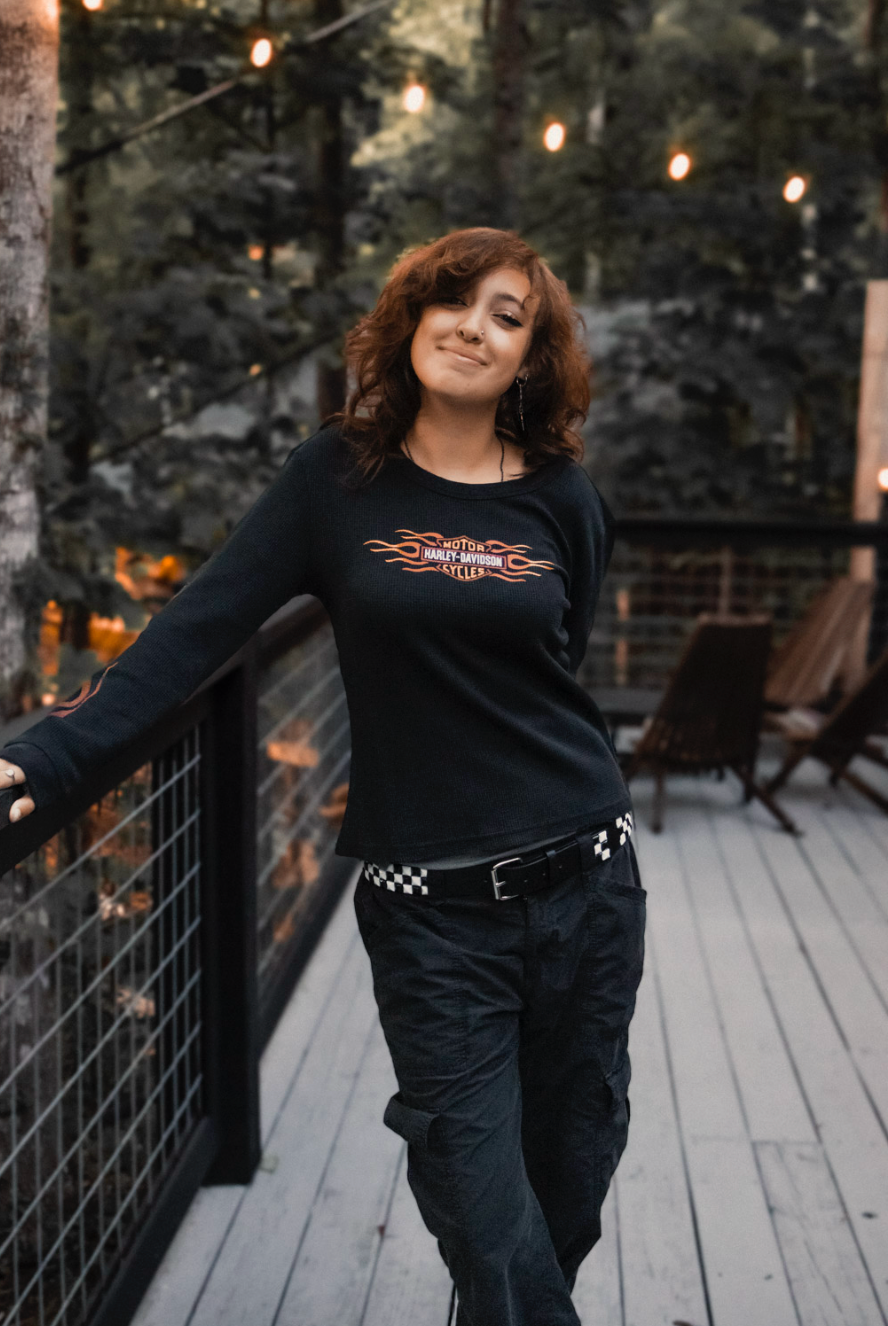 Woman in black outfit dancing and posing playfully on wooden deck at dusk with string lights in background.