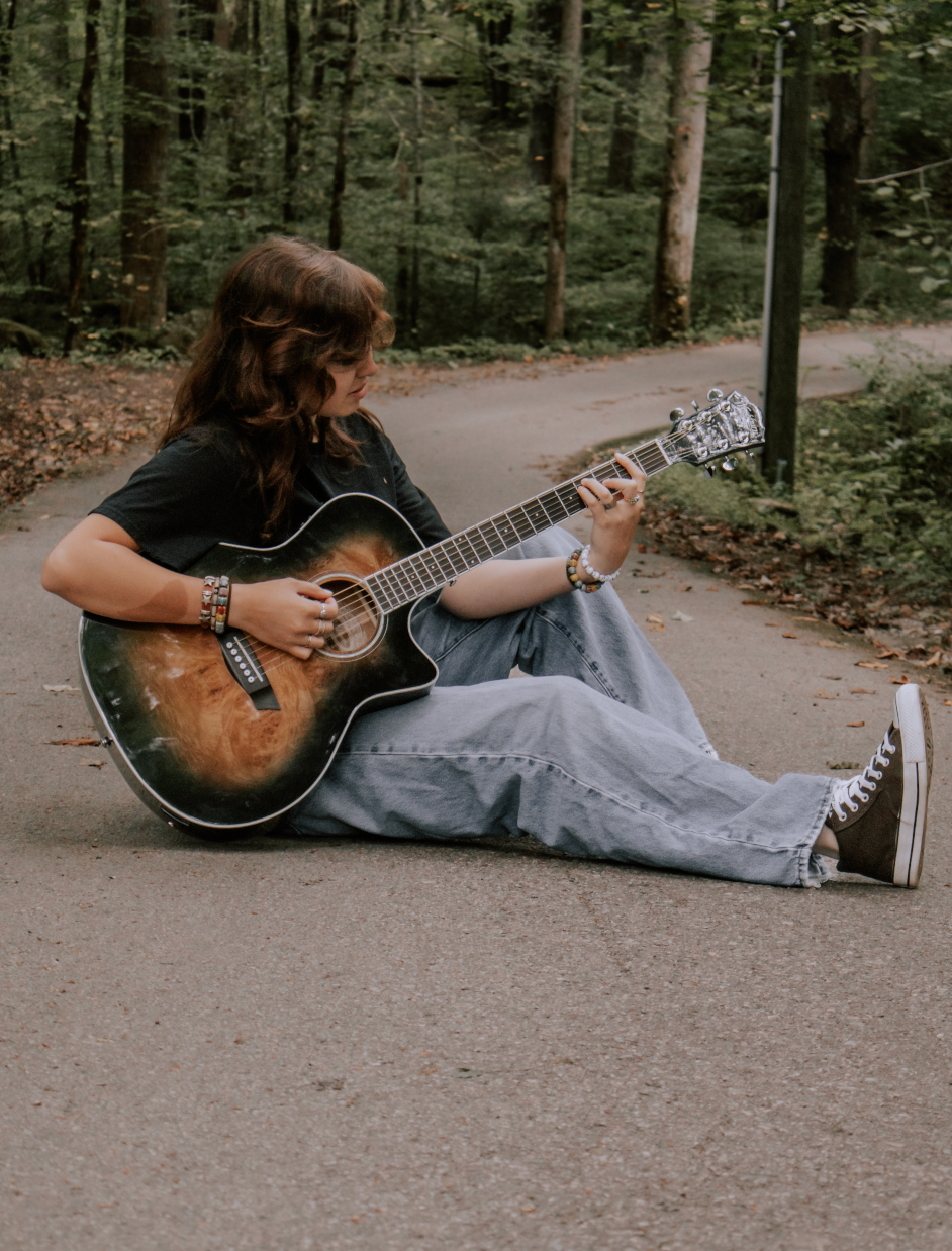 Person lying on concrete path in forest playing acoustic guitar while wearing gray pants and dark top.