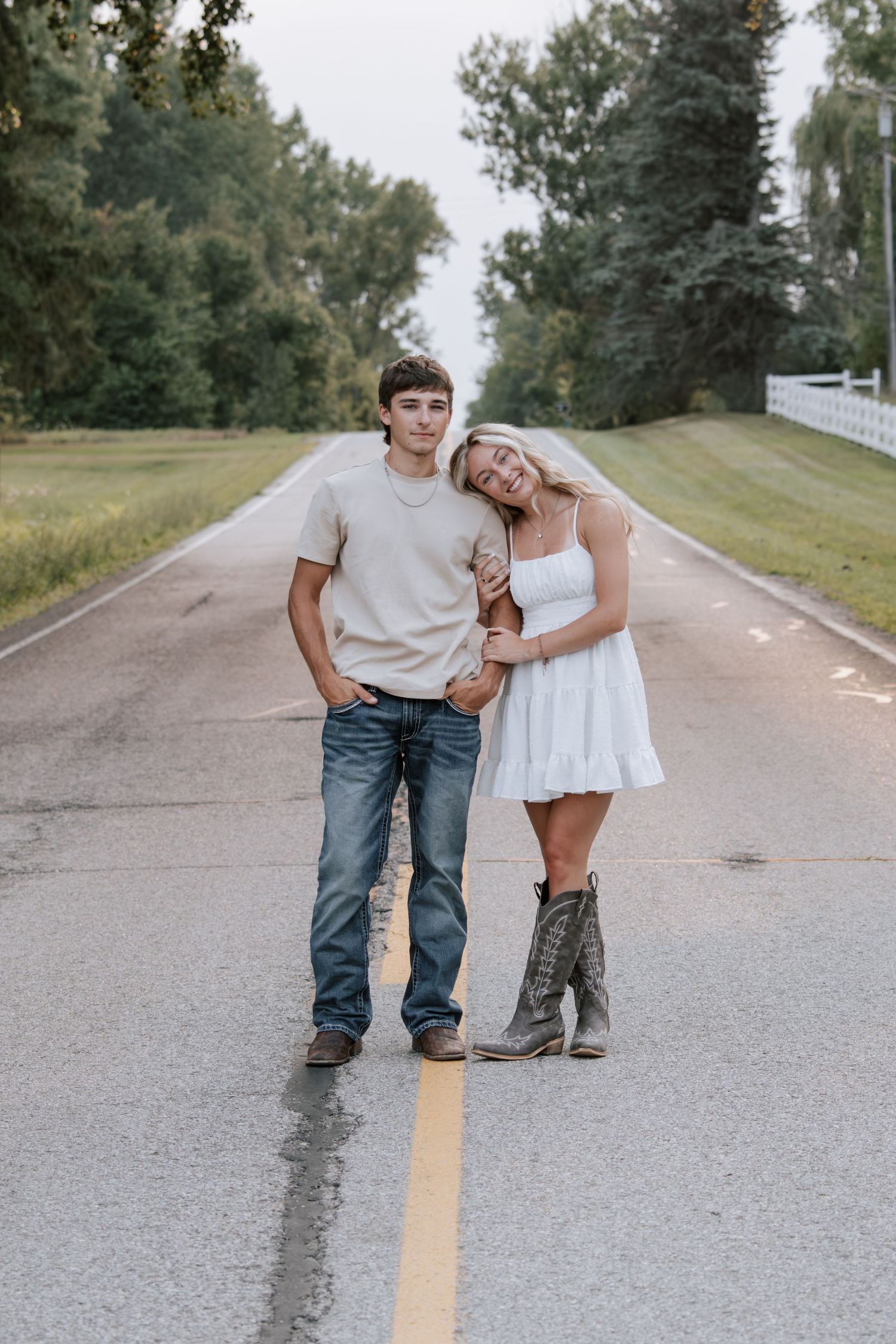 A young couple poses on a rural road, the girl in a white dress and cowboy boots leaning on the boy in jeans.