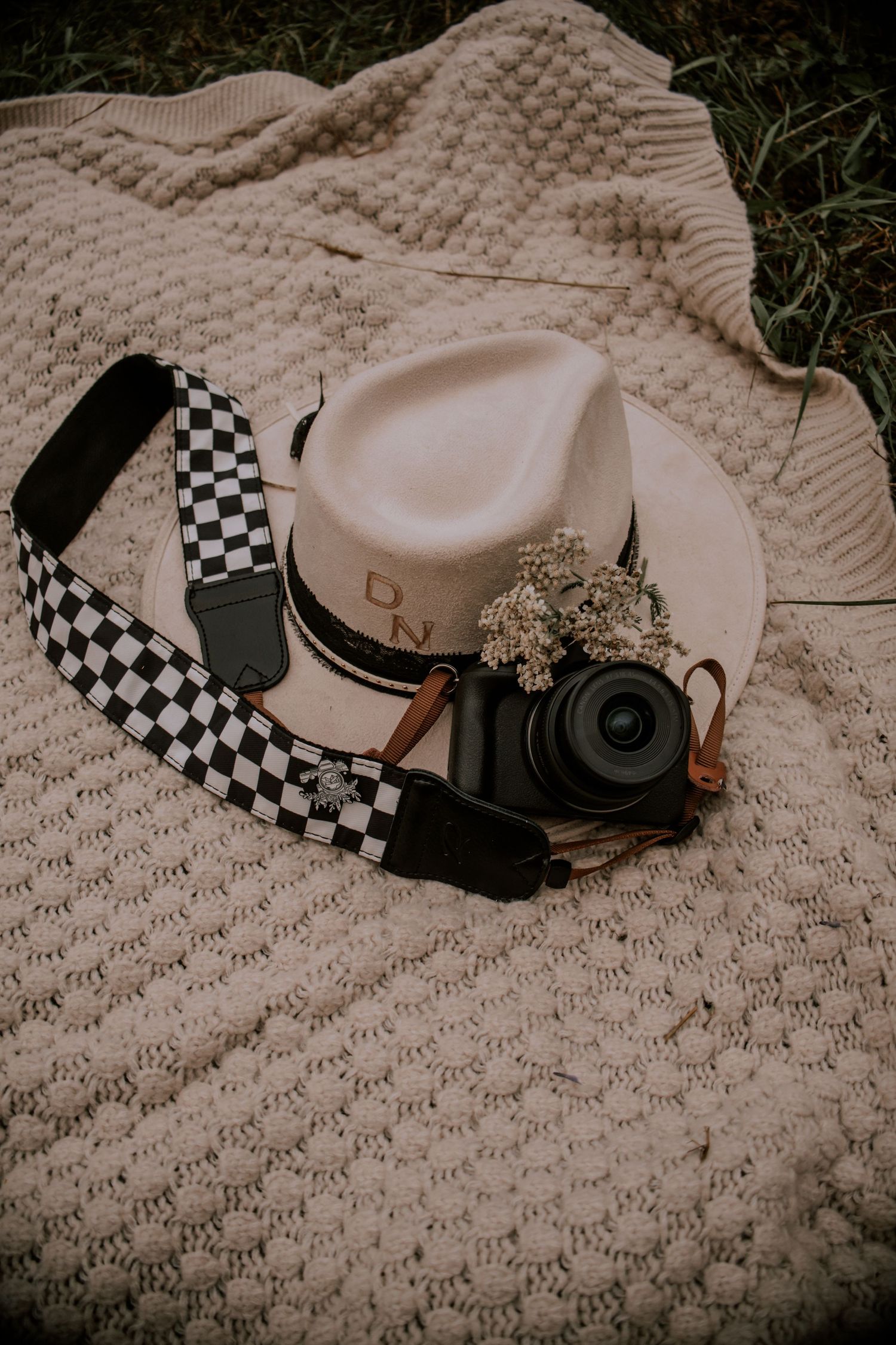 A black camera with checkered strap rests beside a cream felt hat with dried flowers on a knit blanket.