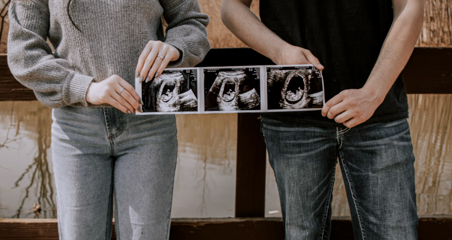 Couple holding ultrasound photos together, announcing pregnancy with sonogram images, wearing casual jeans and sweaters.