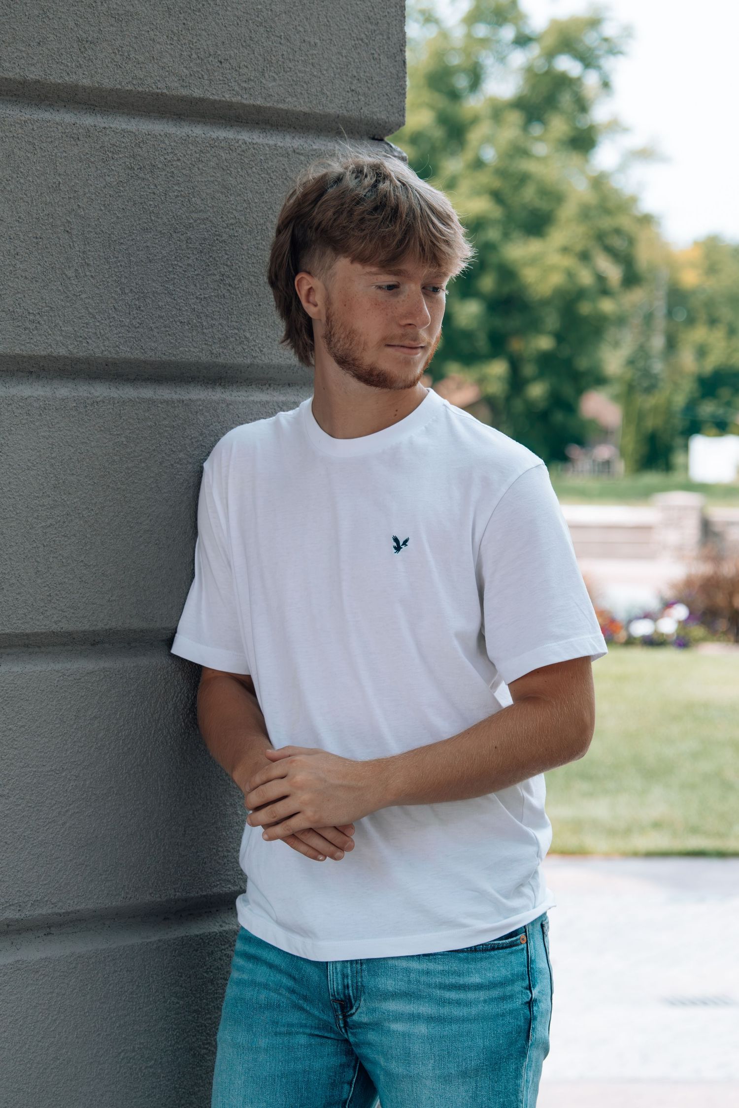 Young man in white t-shirt and blue jeans leaning against a gray wall outdoors.
