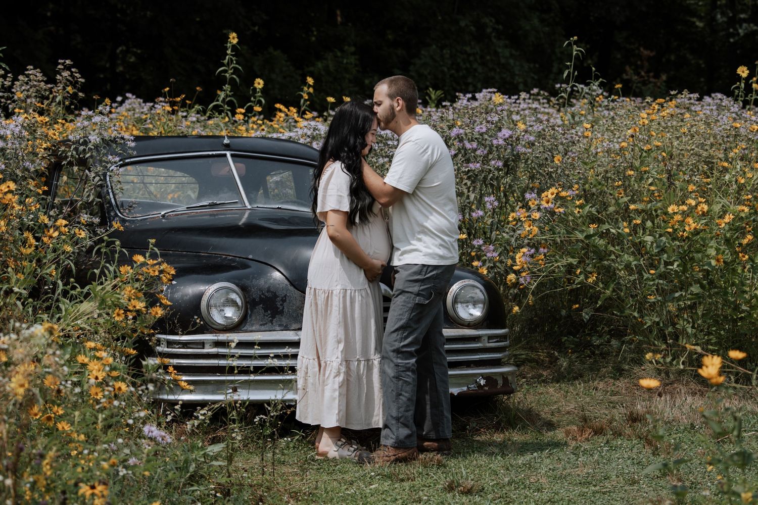 Couple embracing in a wildflower field beside a vintage black car during an outdoor maternity photo session.