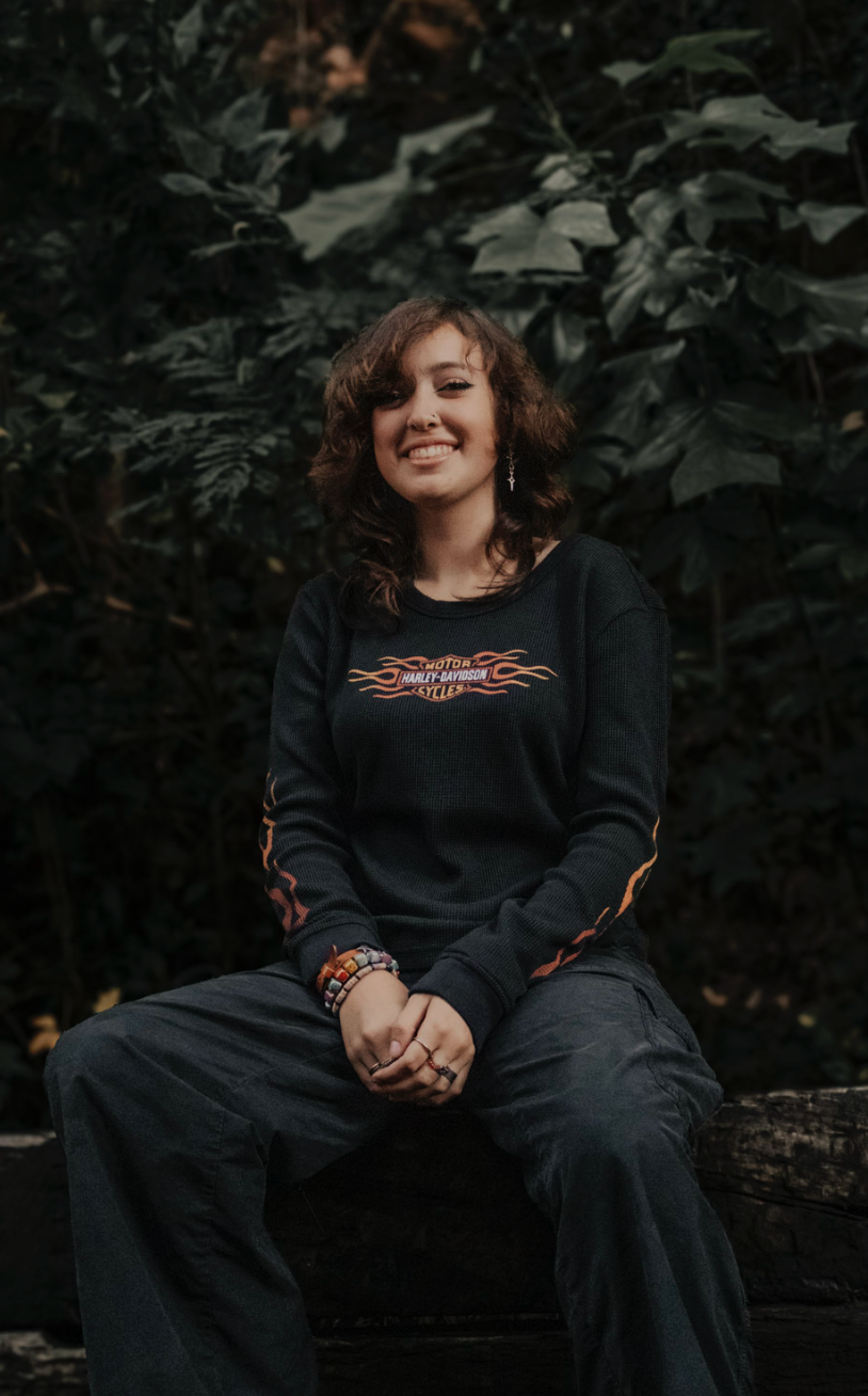 Person in black sweater with orange tribal design sitting on log against dark foliage background in moody outdoor setting.