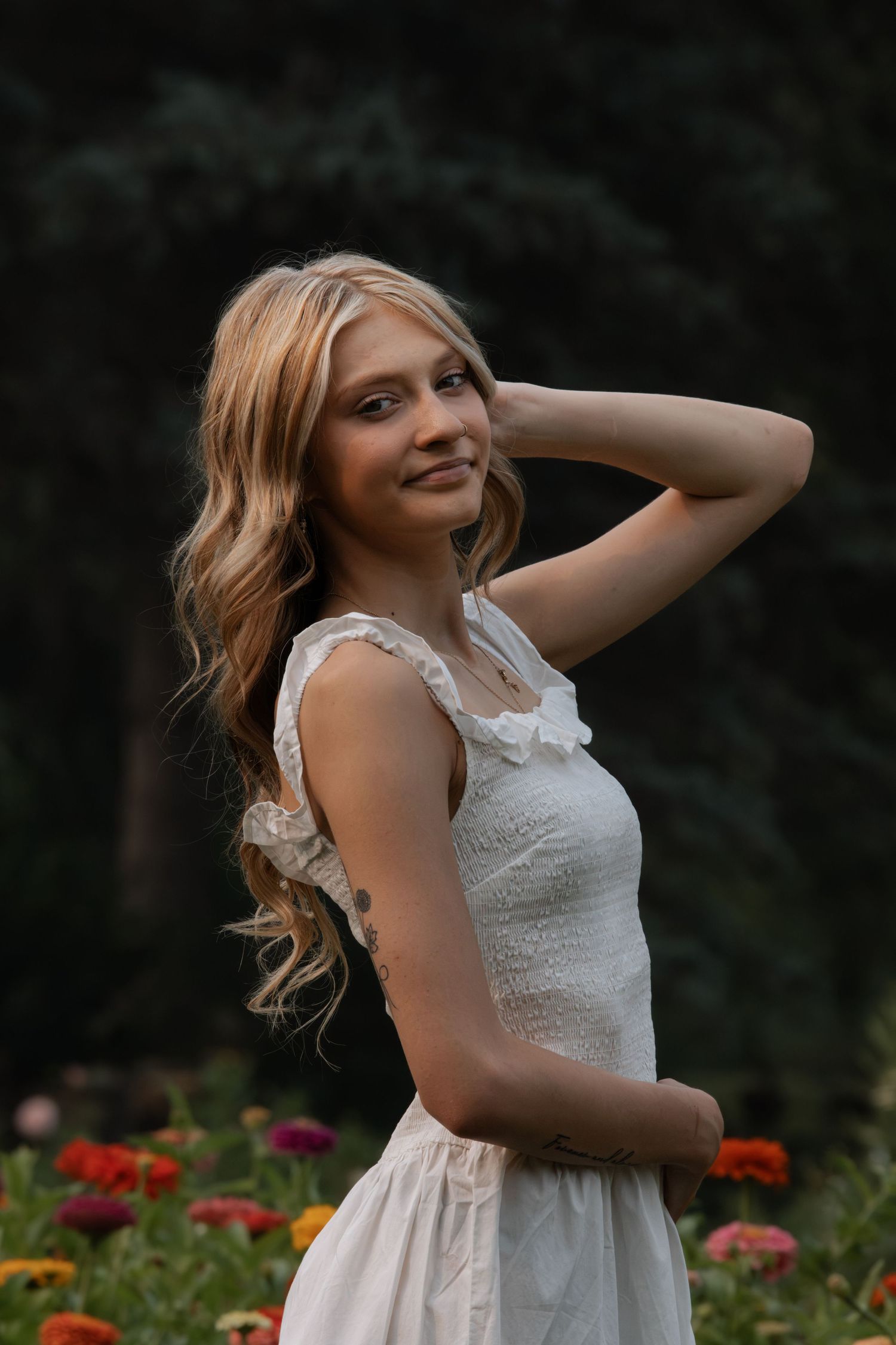 Young blonde woman in white lace dress smiling among colorful flowers in a garden.