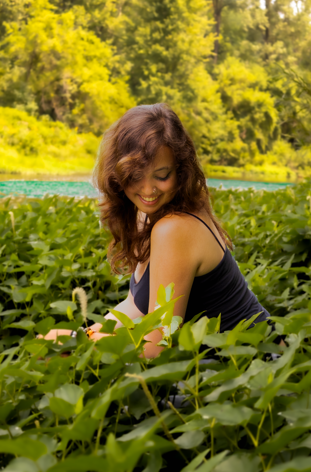 Someone in a black tank top smiles while surrounded by green plants and yellow flowers in nature.