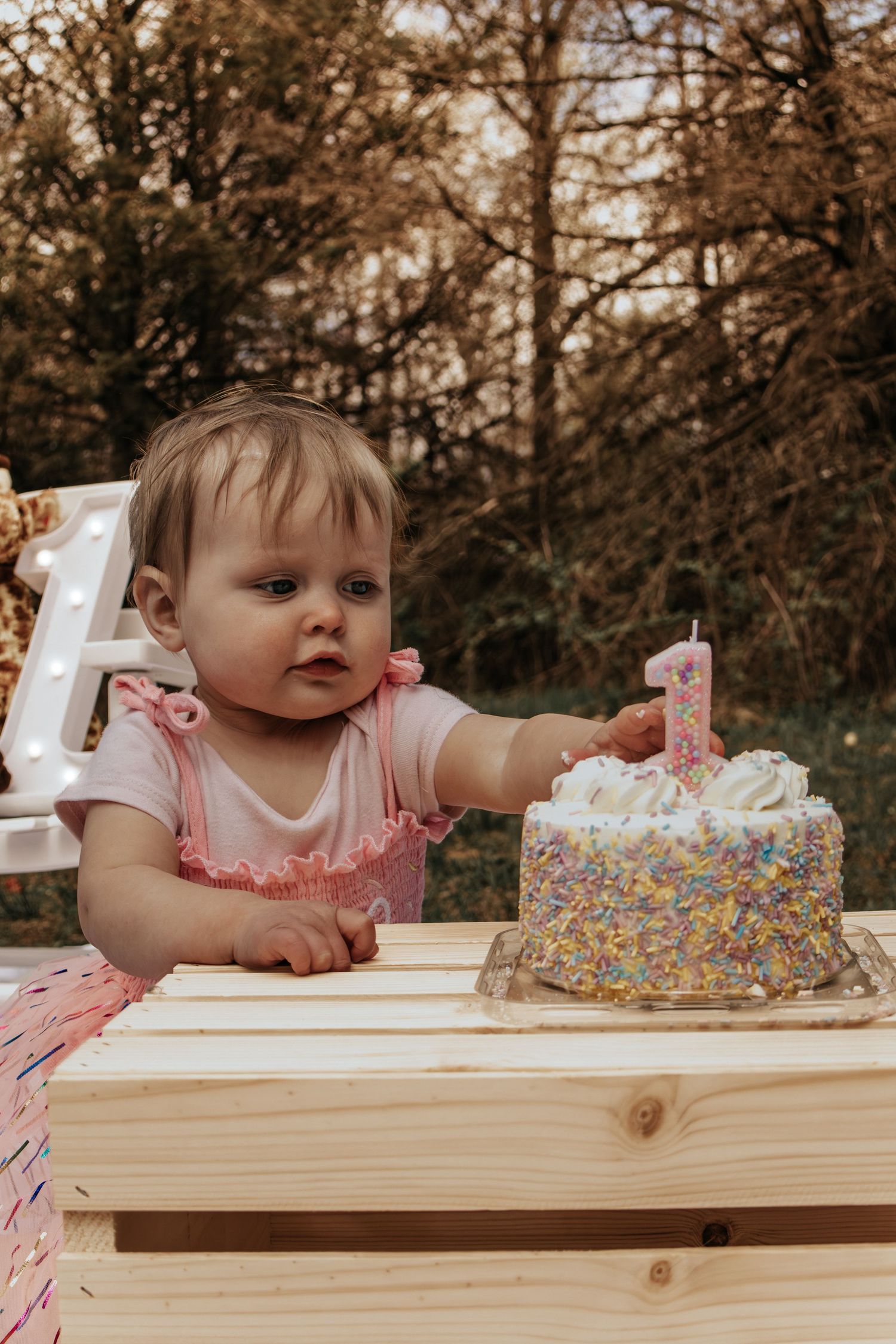Baby girl in pink outfit reaches for her first birthday cake with sprinkles and number one candle outdoors.