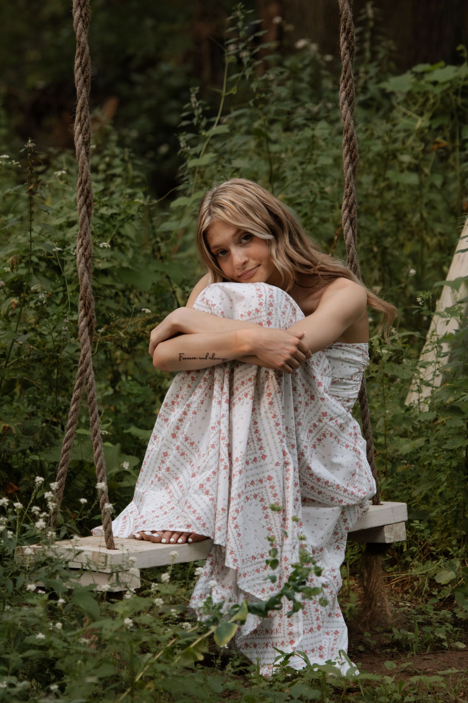 Young woman in floral dress sitting on rope swing surrounded by lush green garden foliage.