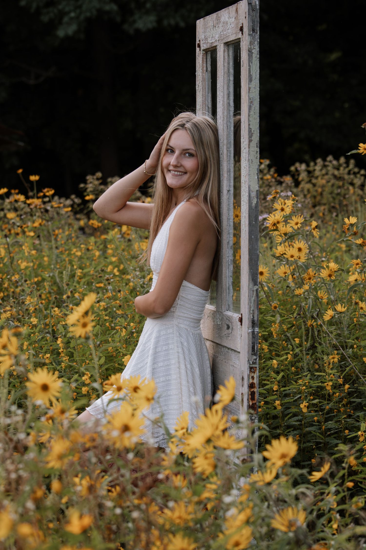Young blonde woman in white dress smiling among yellow wildflowers beside rustic vintage door frame outdoors.