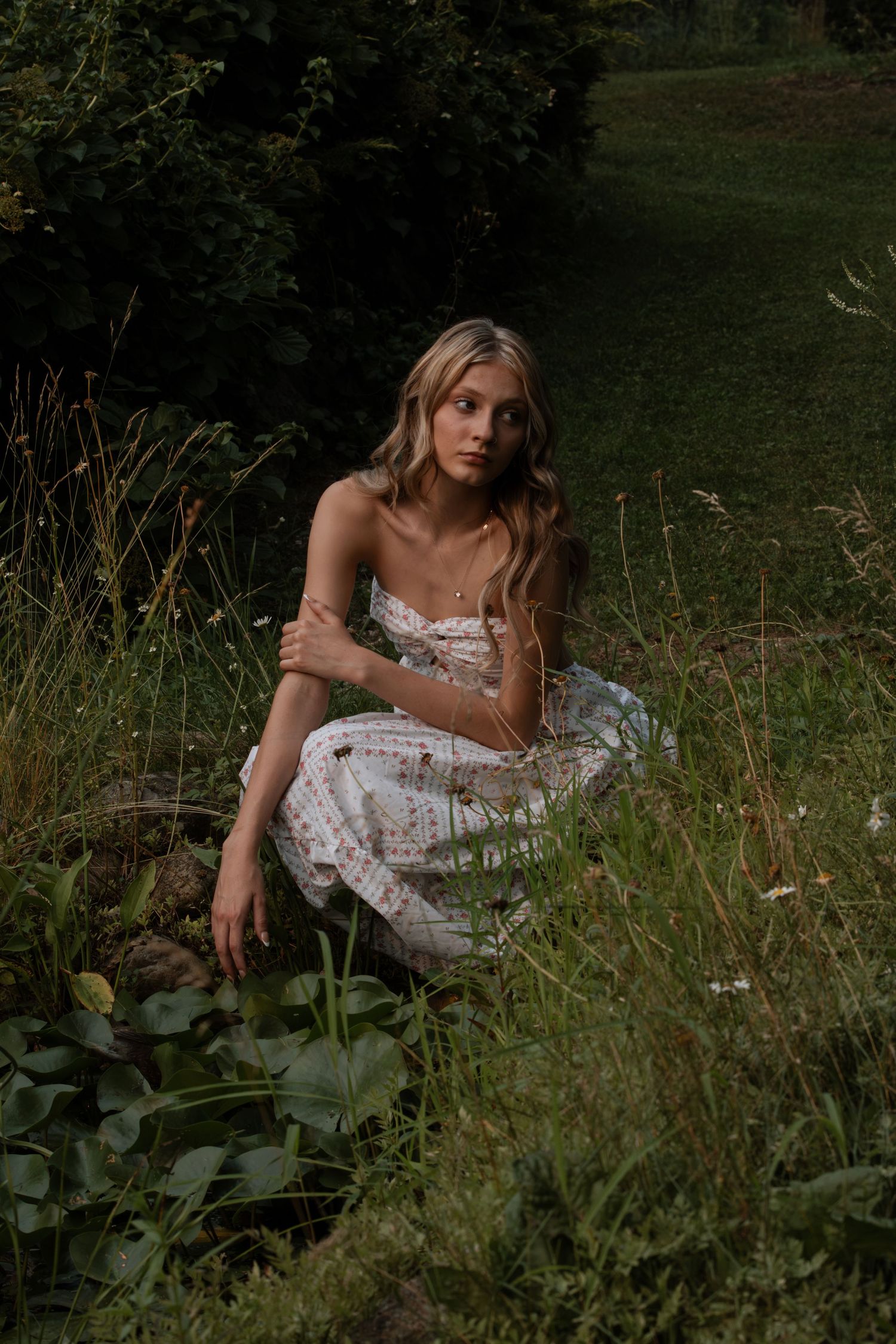 Young woman in white floral sundress sitting gracefully among wild green grass and flowers in a lush garden setting.